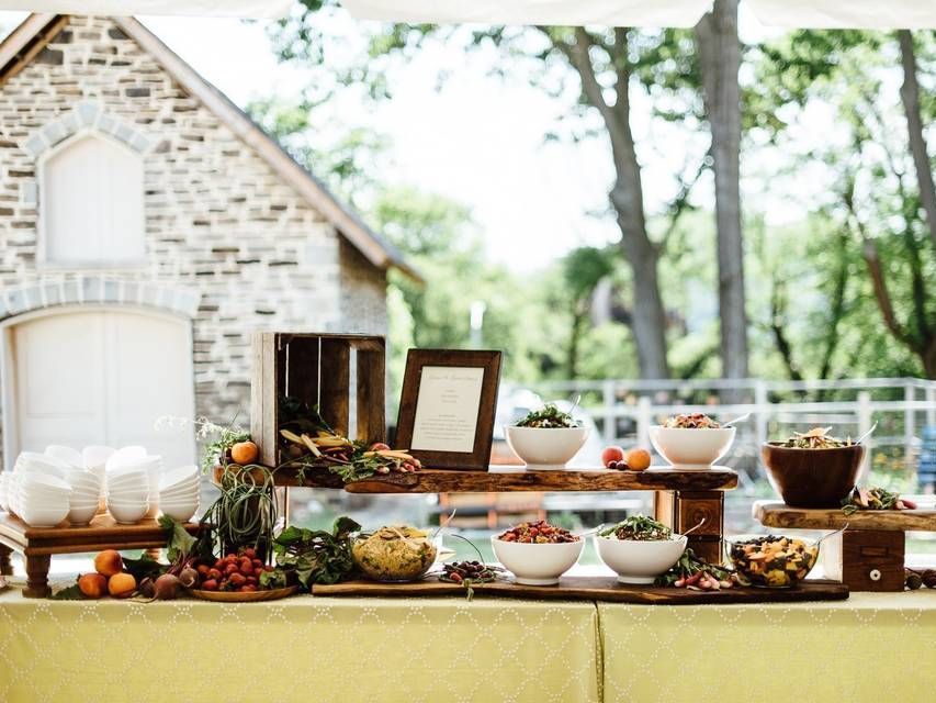 Food buffet table with various salads and fruits set up outdoors in front of a stone building.