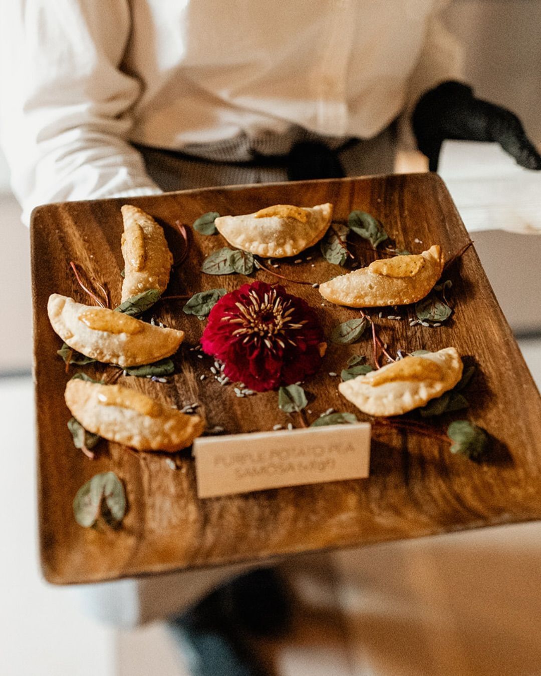 Wooden platter with six pastries, a red flower, and greens, held by a server.