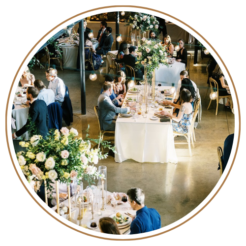Wedding reception with tables set for guests, floral centerpieces, and people dining in a well-lit space.
