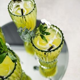 Three green-tinted glasses with yellow liquid and ice cubes, garnished with herbs and a flower, on a silver tray.