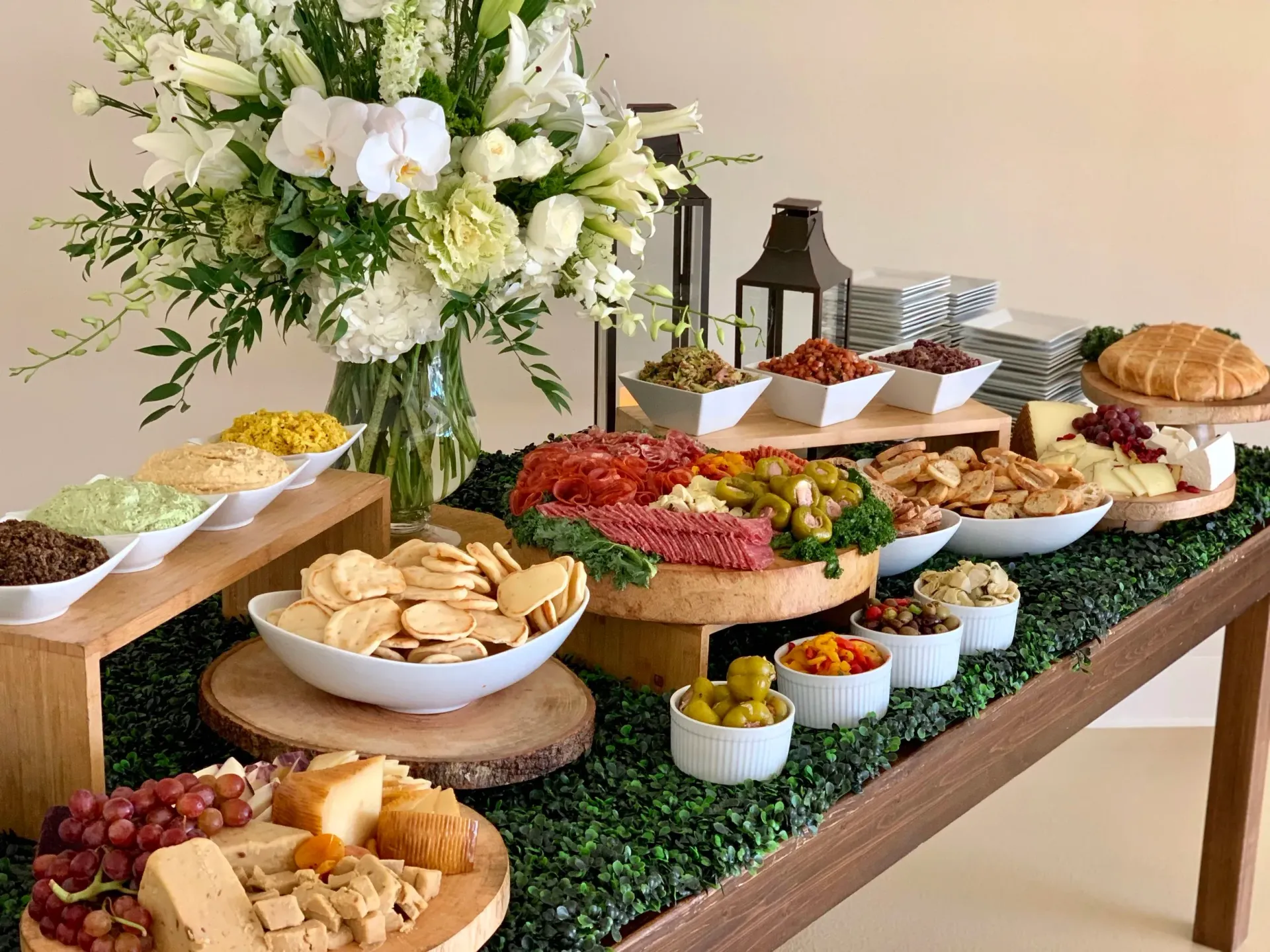 Charcuterie spread on a wood table, with flowers, cheeses, olives, crackers, and dips.