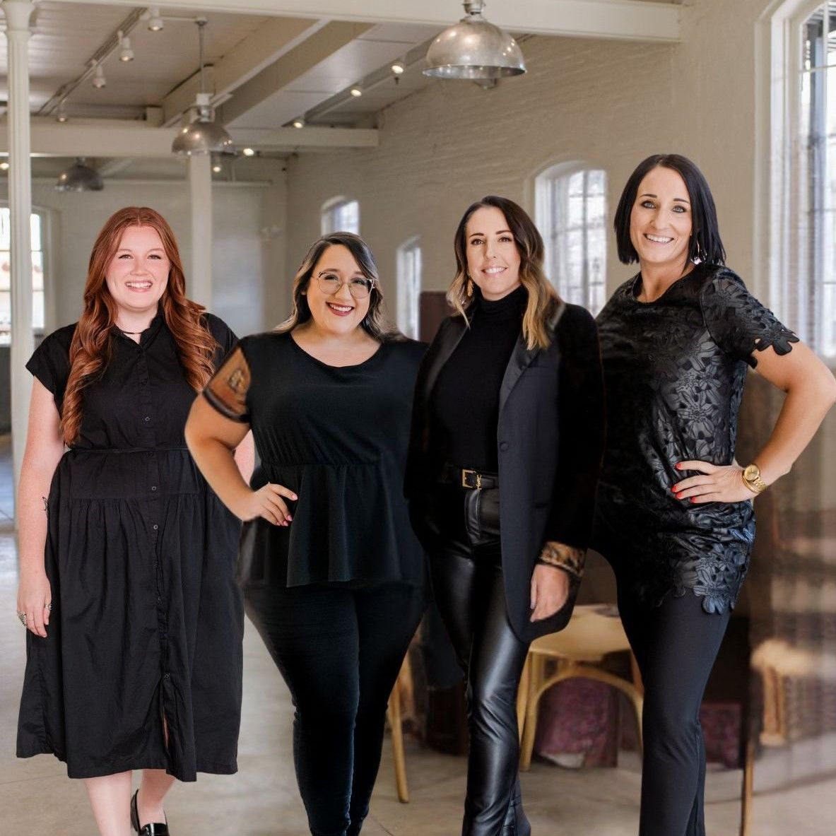 Four women in black outfits pose in a room with tall windows and a high ceiling.