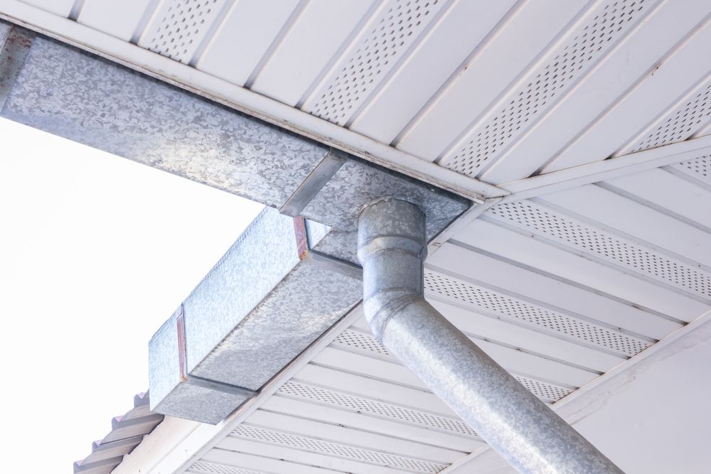 Galvanized metal gutter system on a white-paneled building exterior, showing a horizontal trough connecting to a downspout.
