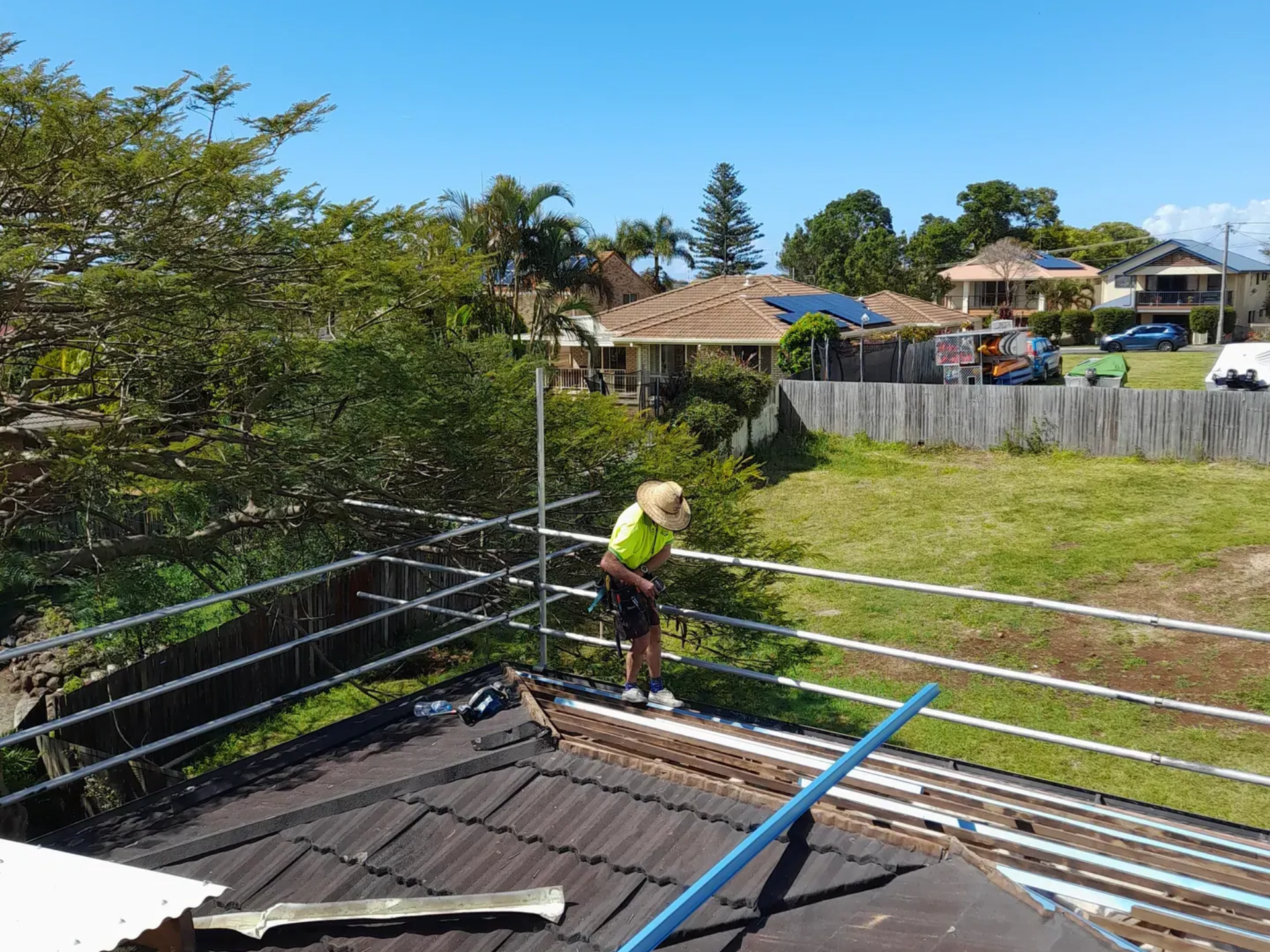 Roofer on a tiled roof with scaffolding, working on a sunny day. Houses and trees in background.