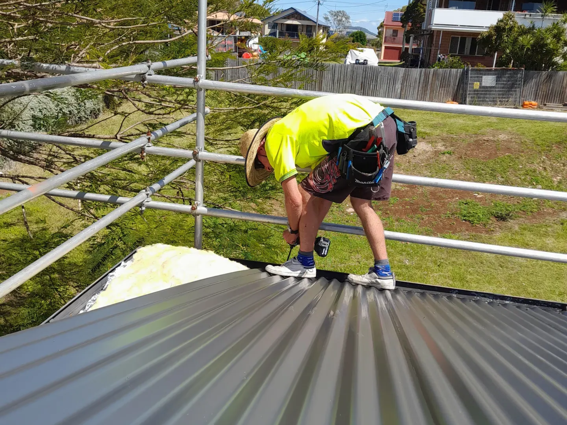 Worker on a metal roof, using a drill. He's wearing a safety vest and is surrounded by scaffolding.