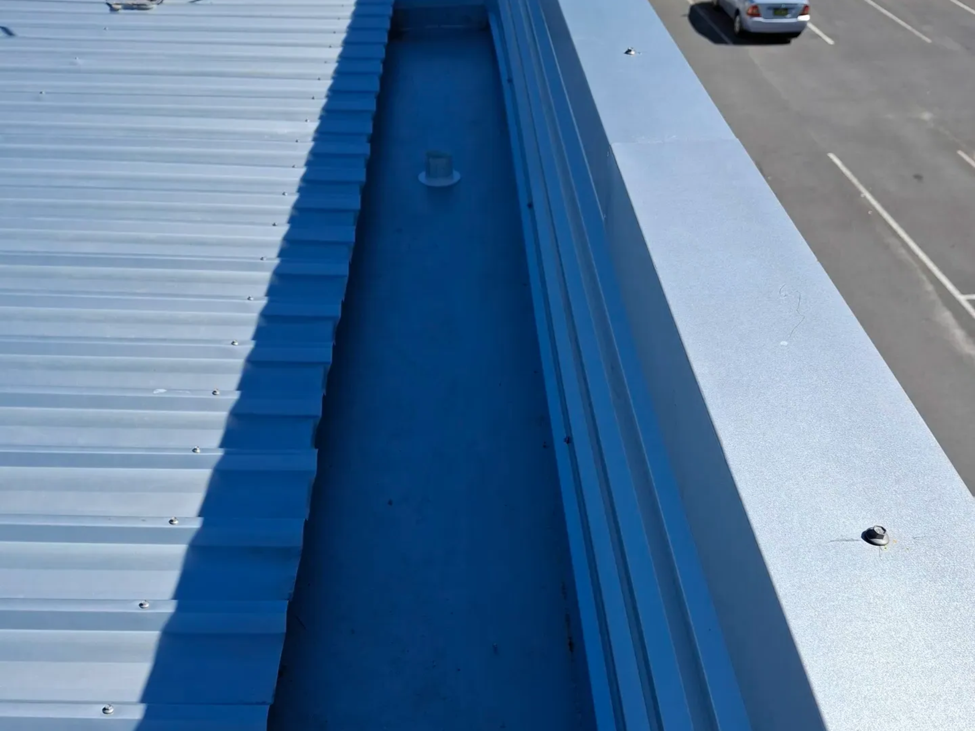 A long, blue metal roof gutter on a building; a car is visible on the adjacent road.