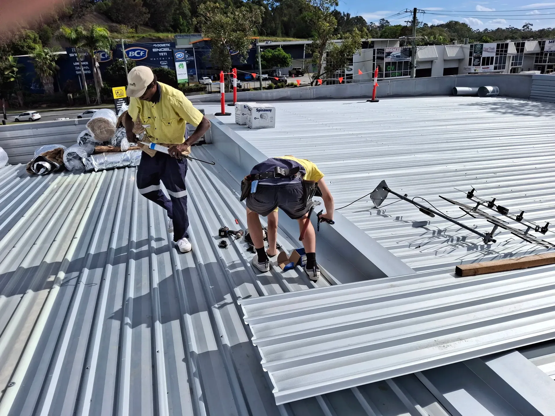 Two workers on a metal roof applying sealant. One is standing, the other is bent over near safety lines.