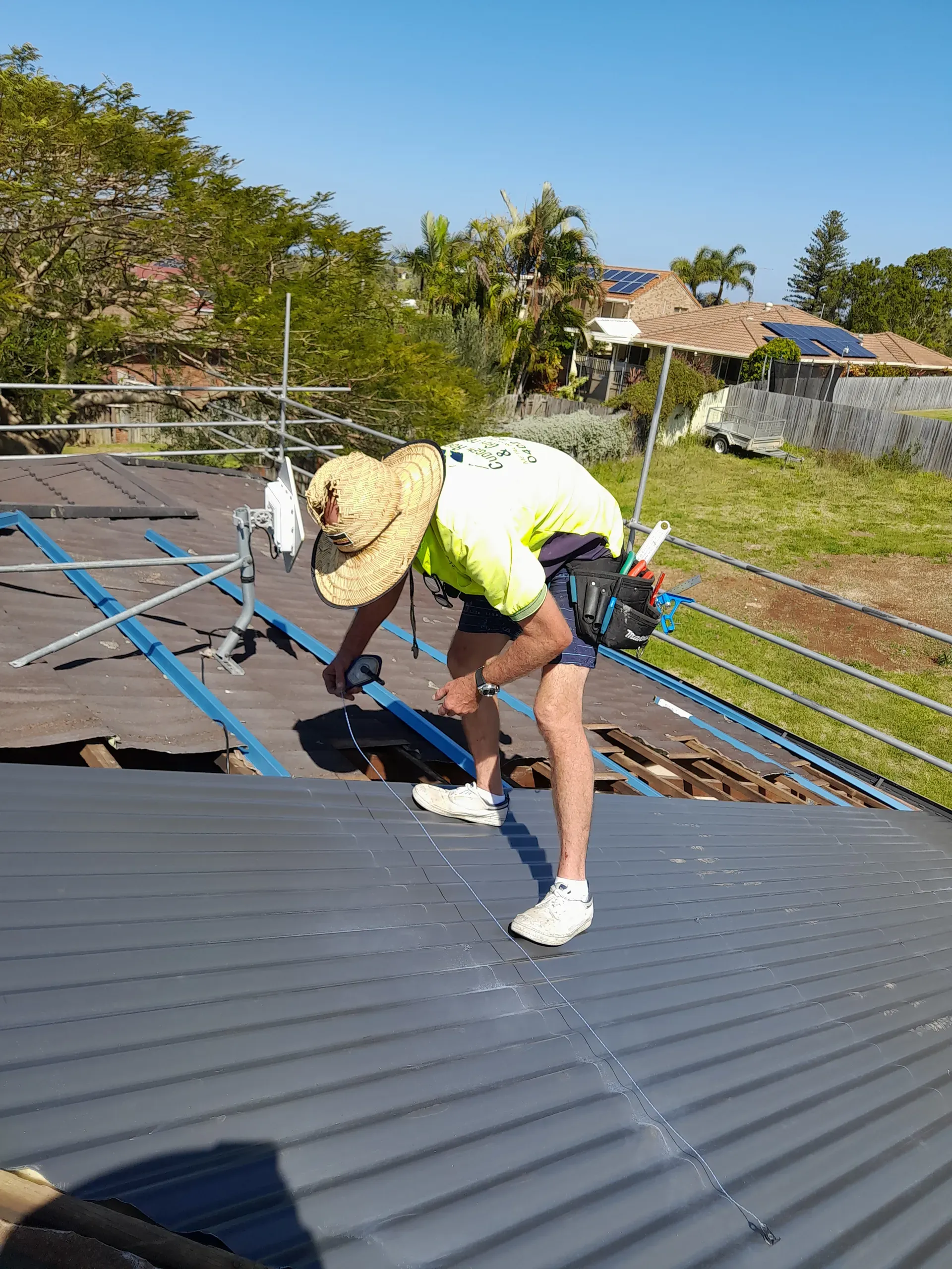 Roofer in safety harness working on a metal roof under blue sky, wearing a sun hat and reflective shirt.
