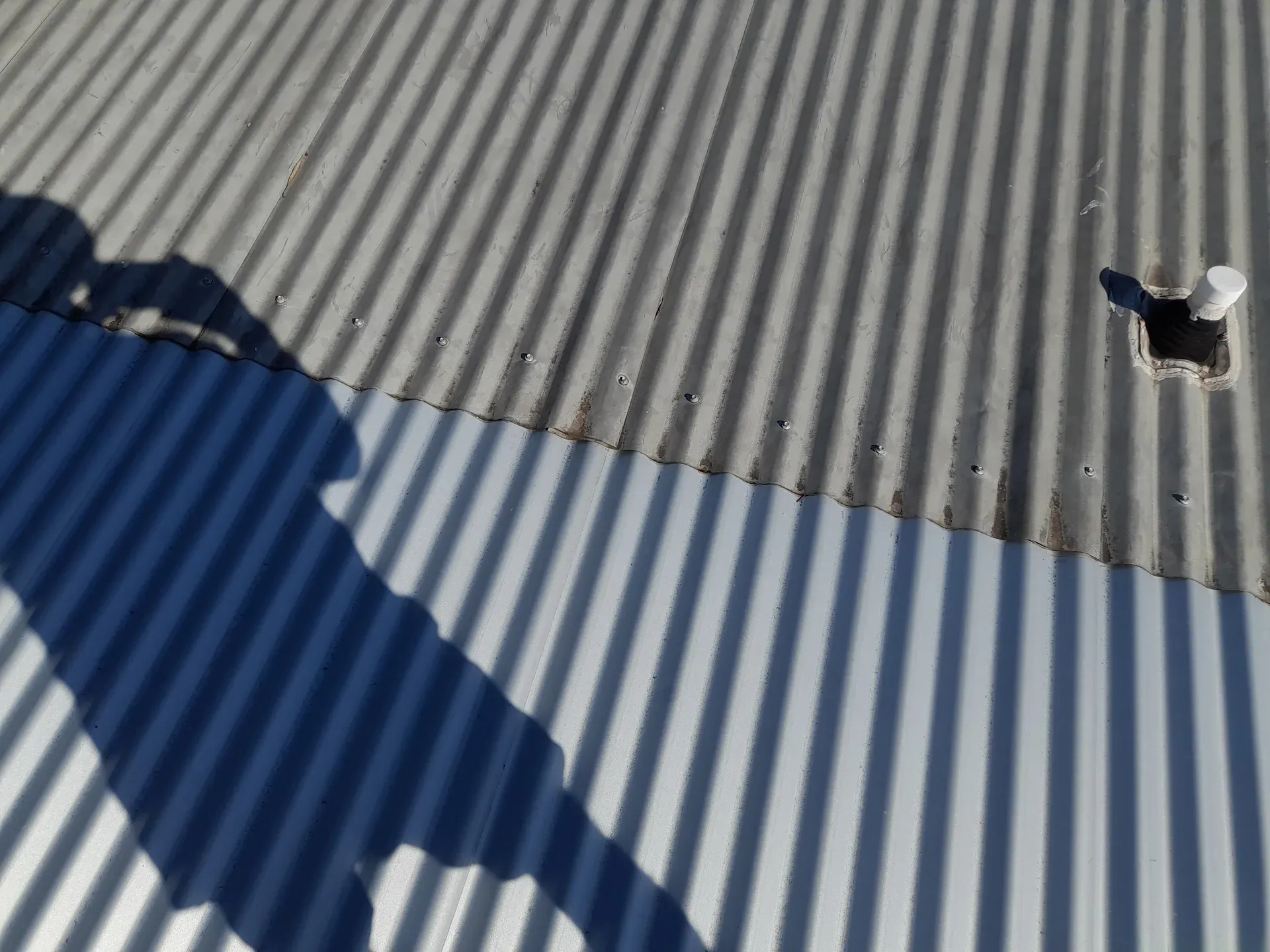 Corrugated metal roof with a shadow cast across it, and a ventilation pipe.