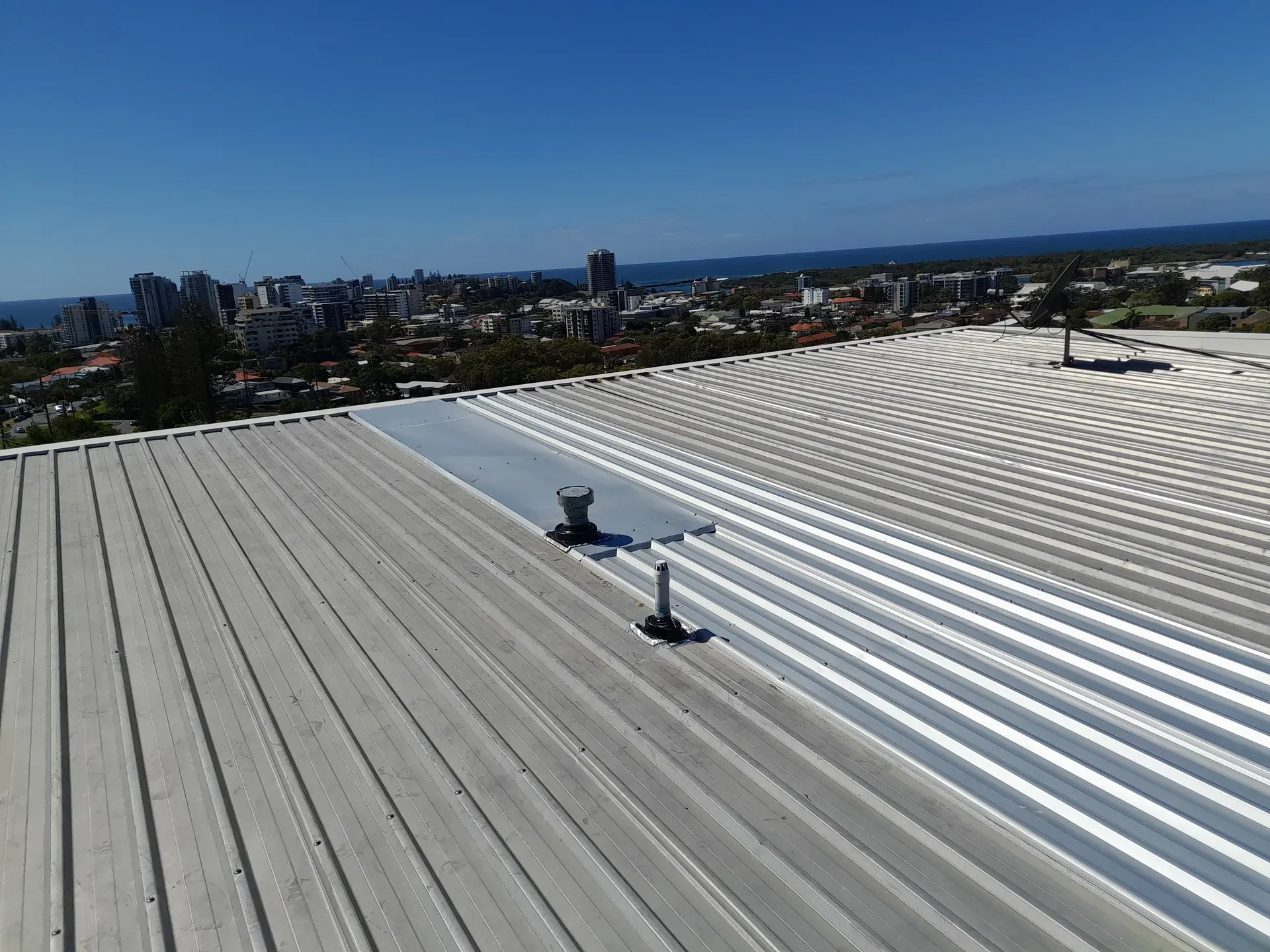 Metal roof with two mounted fixtures, overlooking a city on a sunny day.
