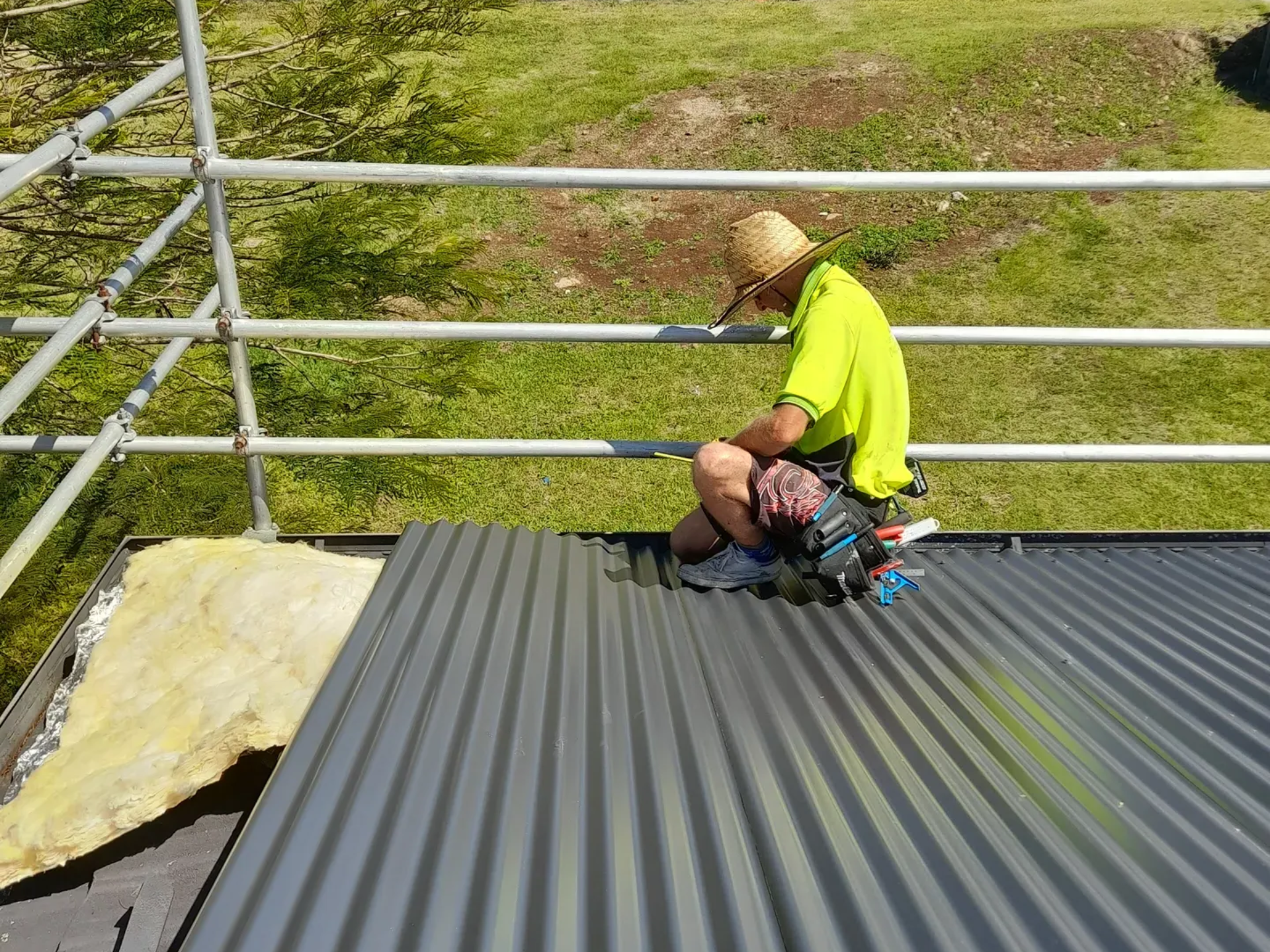 Worker on a corrugated metal roof wearing safety gear, working near scaffolding and insulation, outdoors.