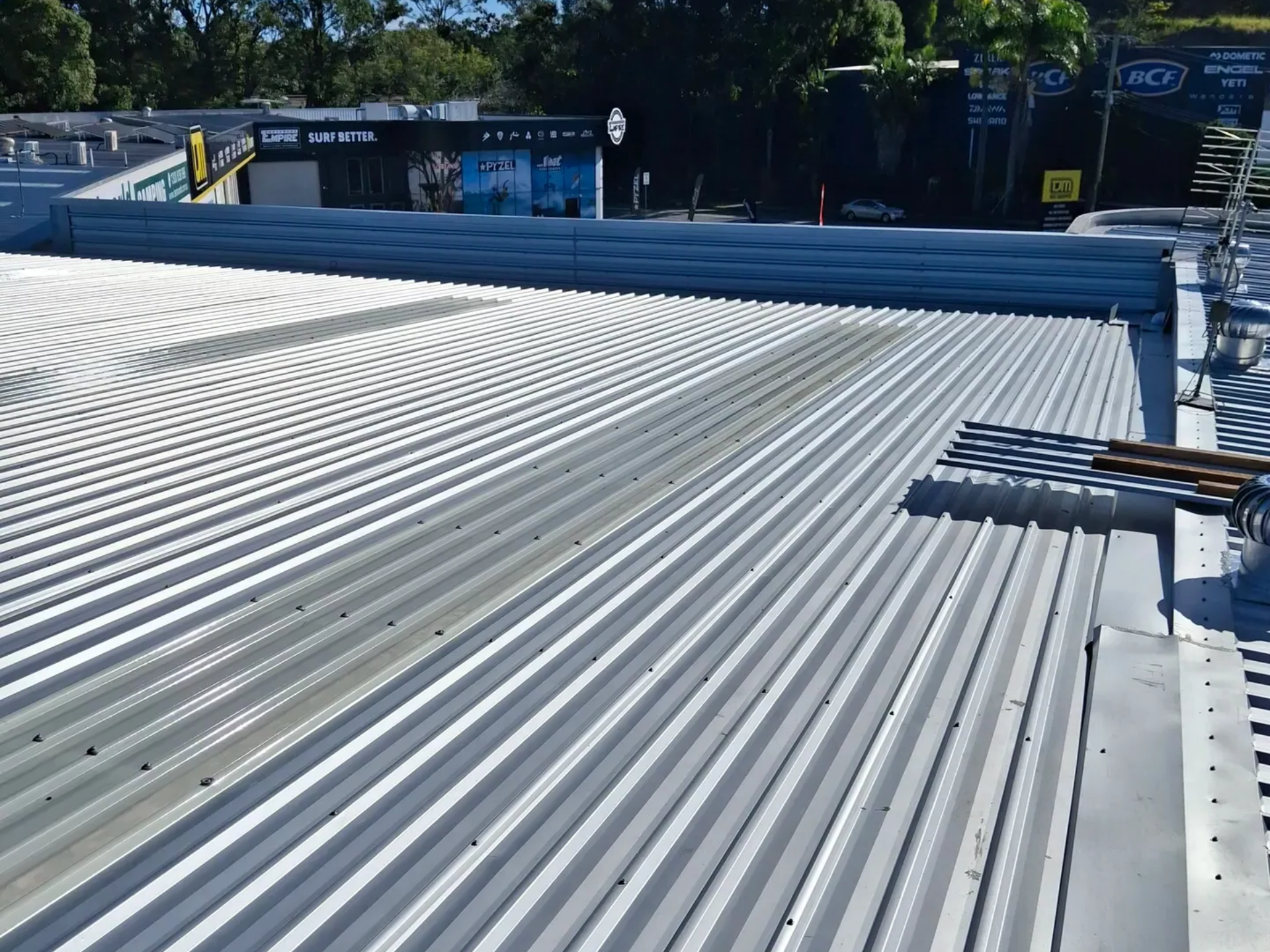 Person in neon shirt using a drill on a corrugated metal roof, with scaffolding and greenery in the background.