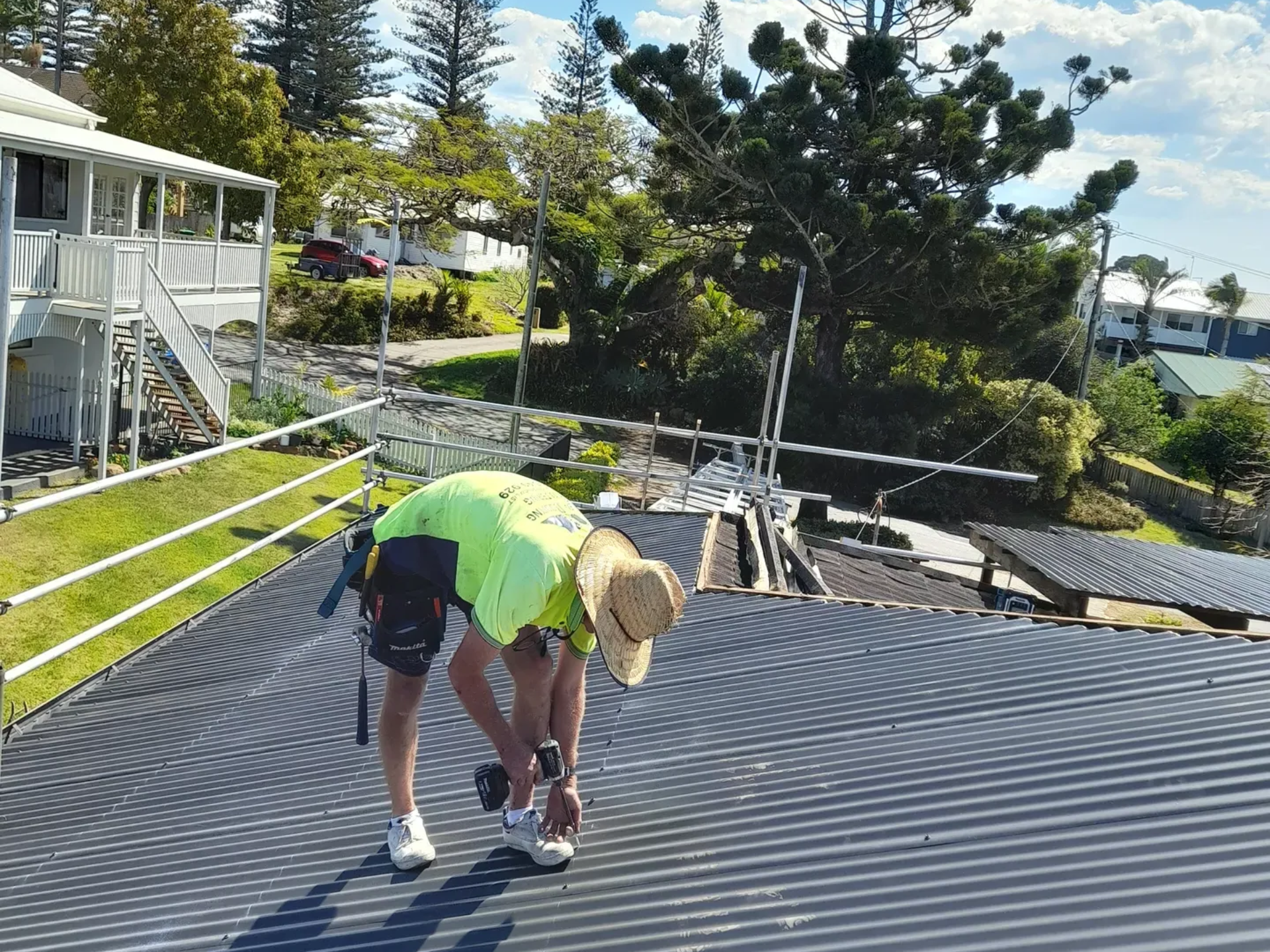 Roofer in safety gear on corrugated roof, working in sunlight, with coastal background.