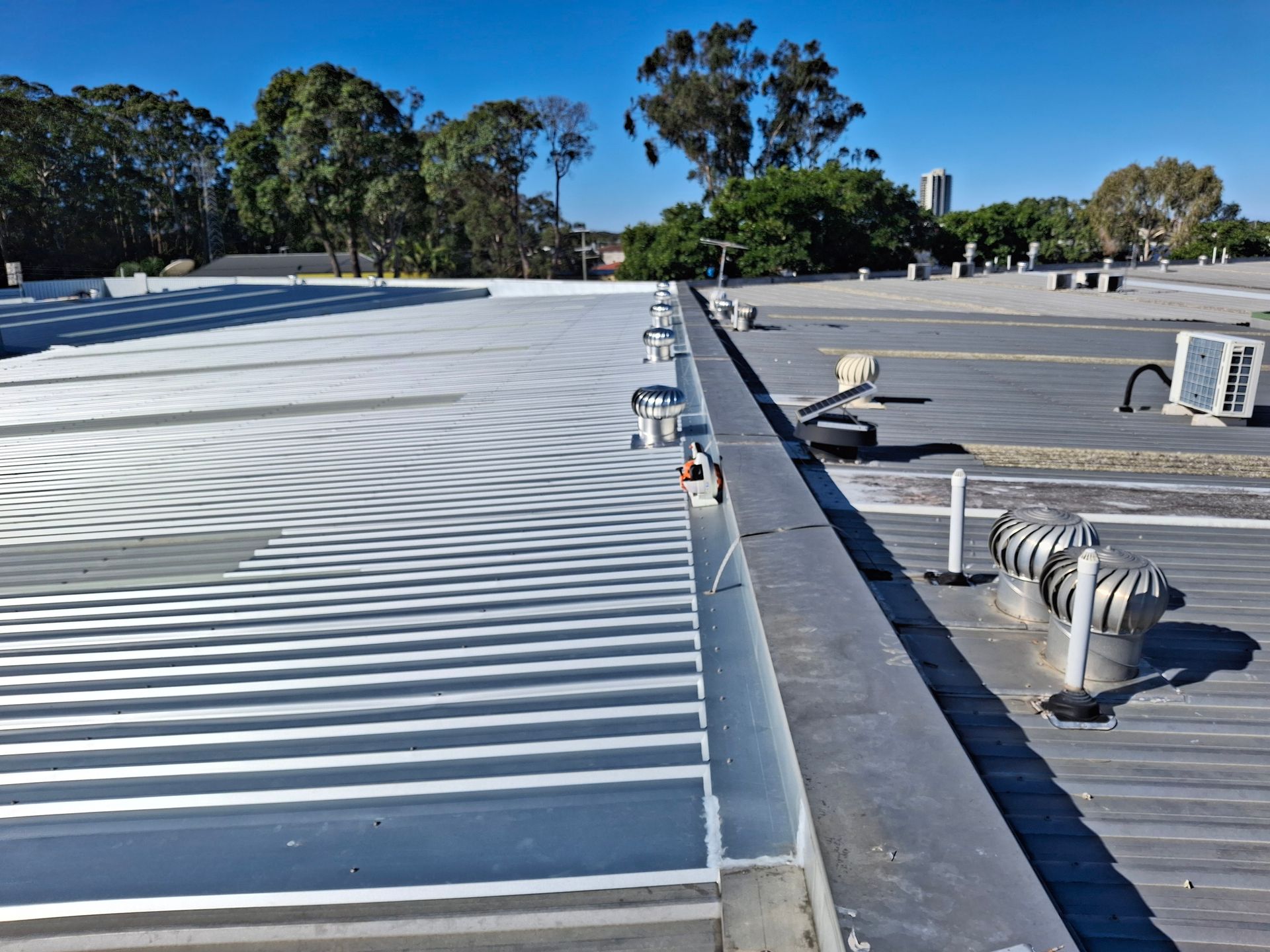 Metal roof with air conditioning units and vents against a blue sky, trees in the background.