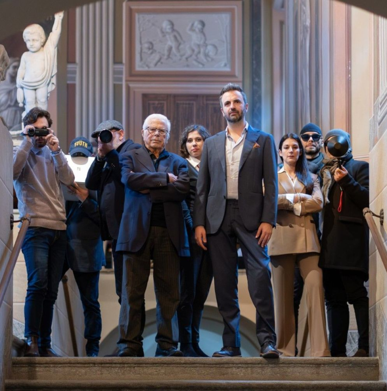 Un gruppo di persone sulle scale di un edificio. Si distinguono uomini in giacca e cravatta. Altri indossano occhiali da sole, cappelli e tengono in mano macchine fotografiche.