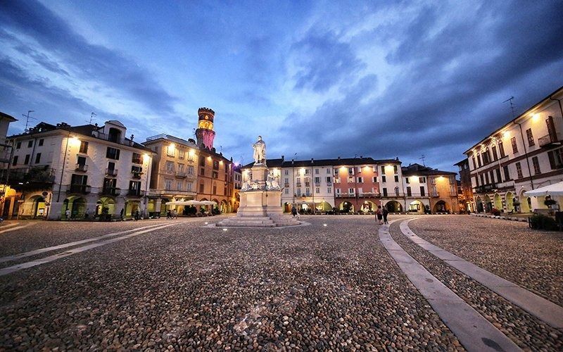 Piazza acciottolata al tramonto; edifici con finestre illuminate, monumento centrale, cielo nuvoloso.