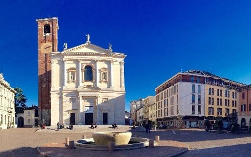 Chiesa e piazza con una fontana di fronte, giornata di sole, cielo azzurro, edifici sullo sfondo.