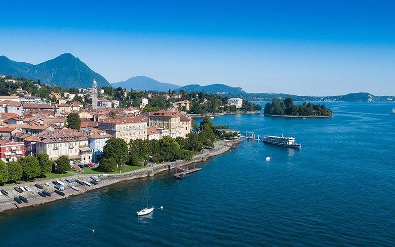 Città sul lago con edifici, barche e una piccola isola. Montagne sullo sfondo, acqua azzurra, giornata di sole.