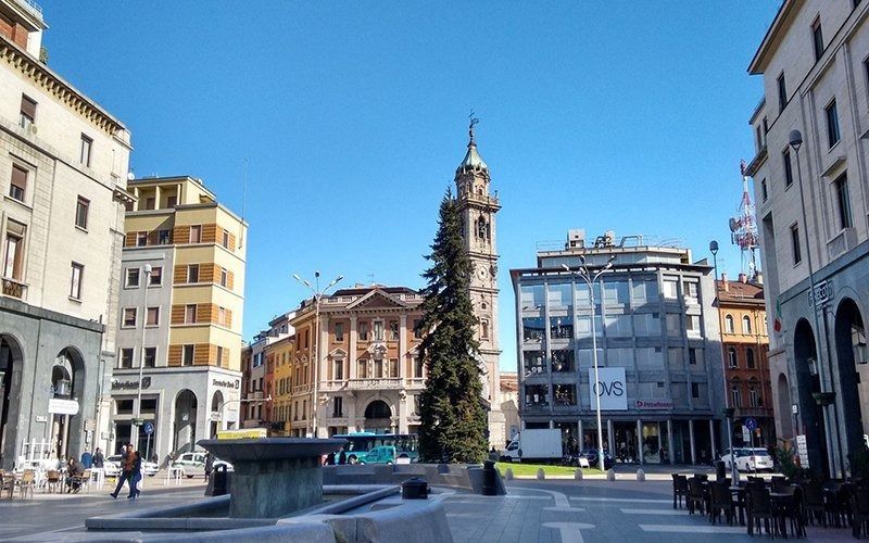 Piazza cittadina con edifici, fontana e alta torre dell'orologio sotto il cielo azzurro.