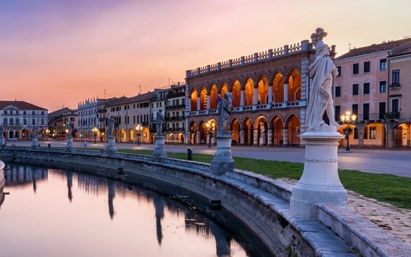 Lungomare lastricato al tramonto. Statue costeggiano un muro di pietra con edifici e archi, riflettendosi nell'acqua.