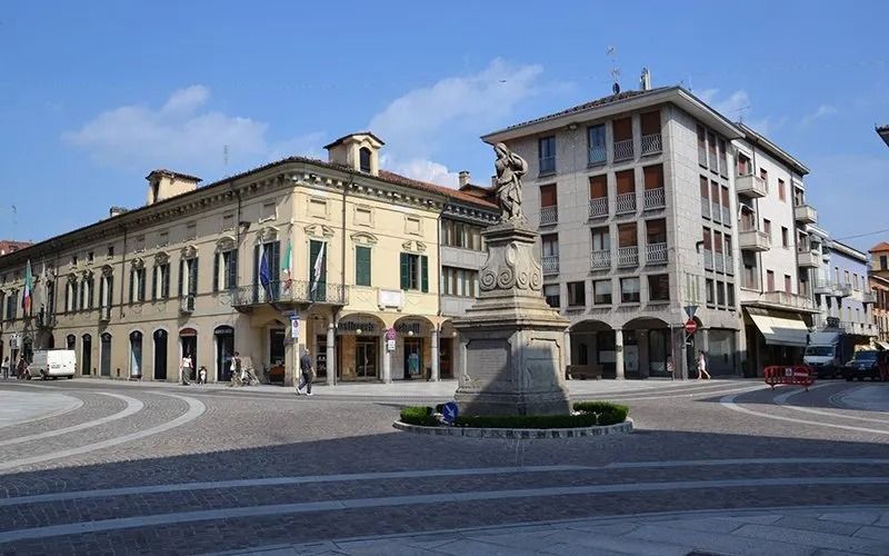 Piazza con edifici, statue e pedoni sotto un cielo azzurro.