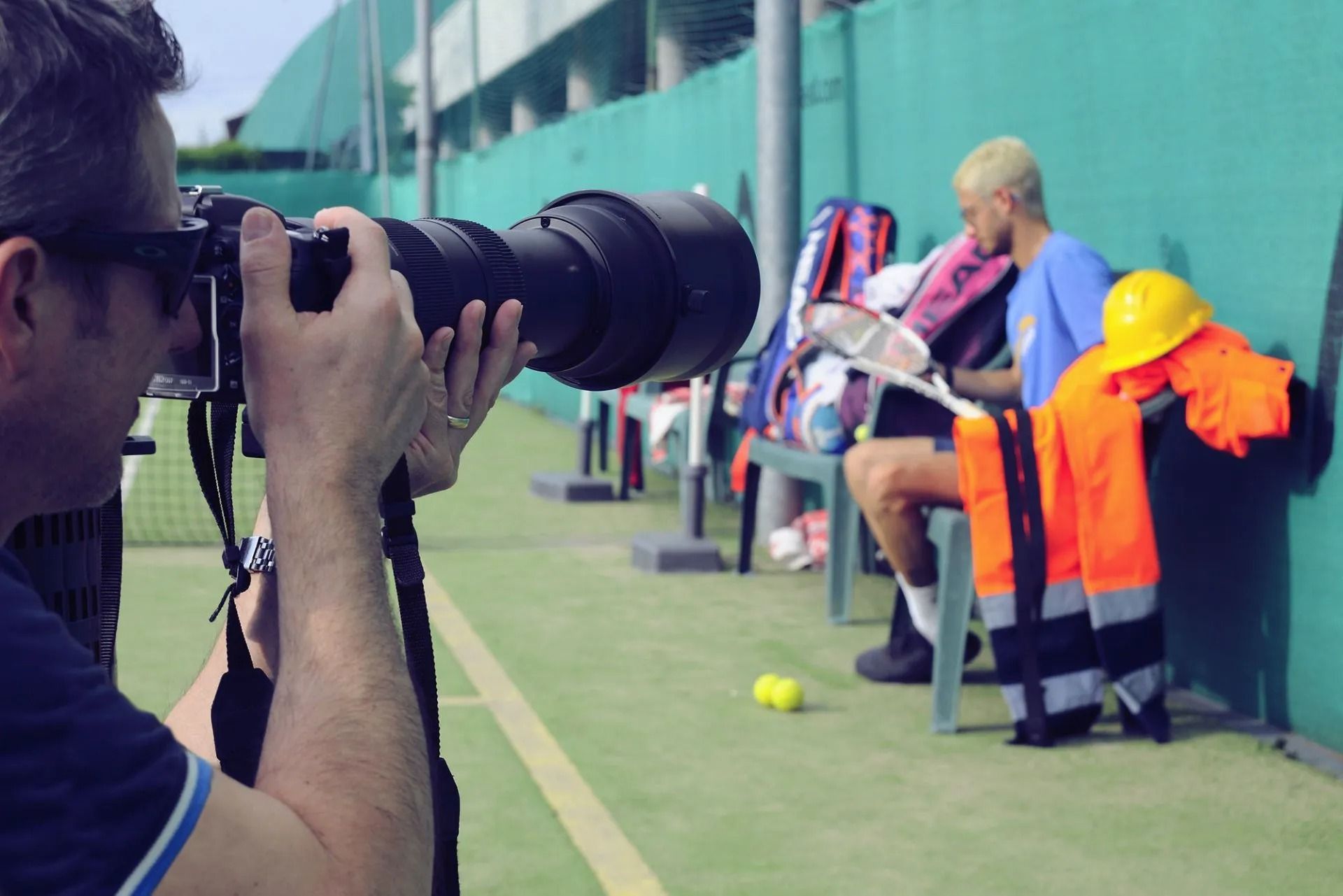 Un fotografo scatta una foto a un tennista che si riposa su una panchina vicino a un campo. Un giubbotto di sicurezza arancione è appeso lì vicino.