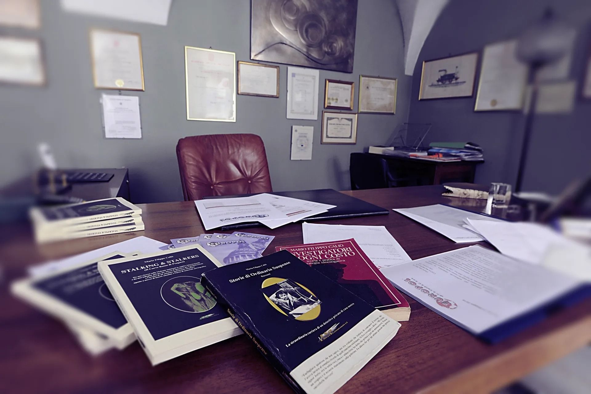 Una scrivania da ufficio con libri, documenti e una sedia in pelle; sullo sfondo, dei diplomi appesi alla parete.