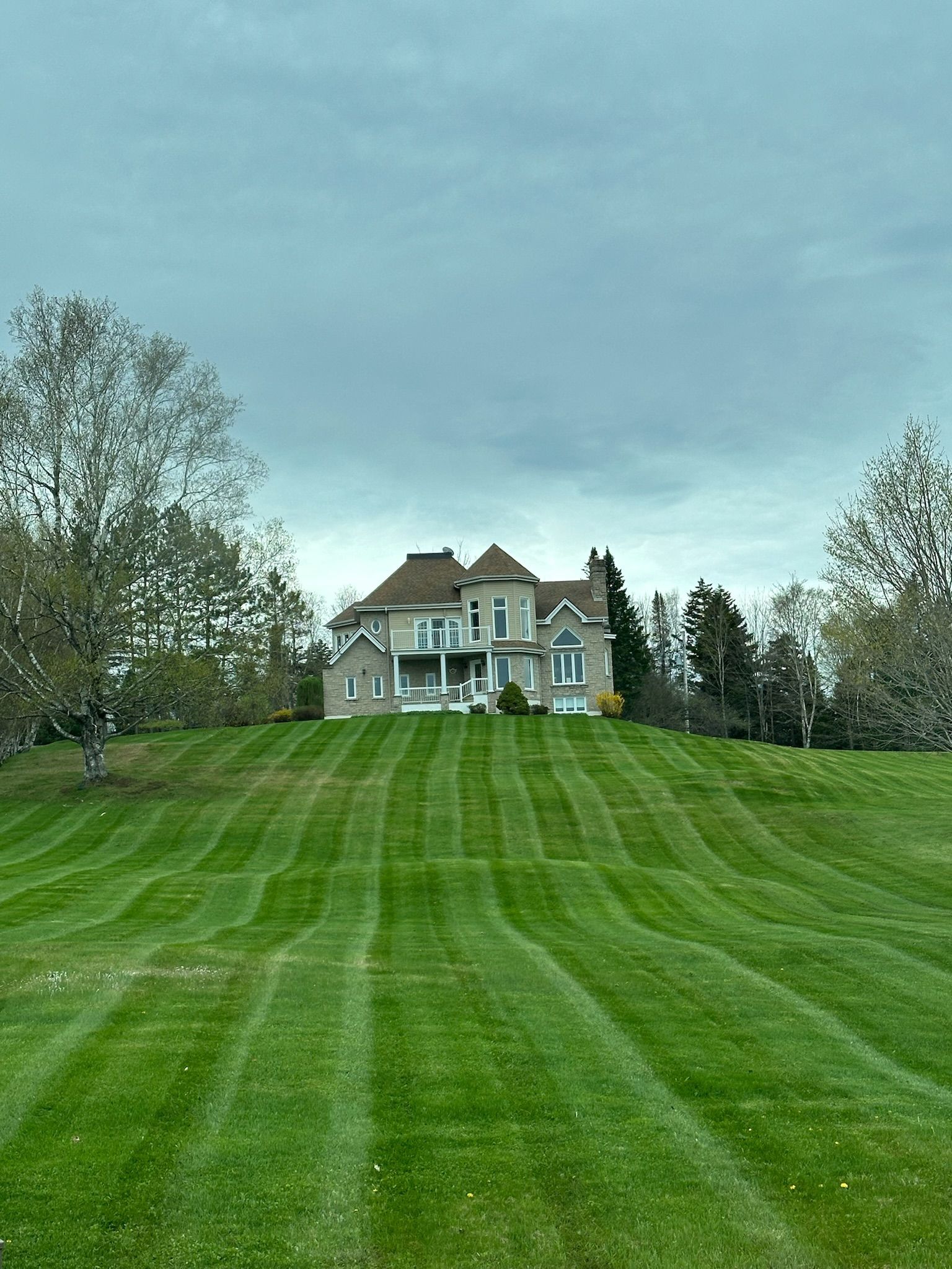 Grande maison blanche sur une colline herbeuse sous un ciel nuageux, vue depuis une pelouse tondue bordée d'arbres.