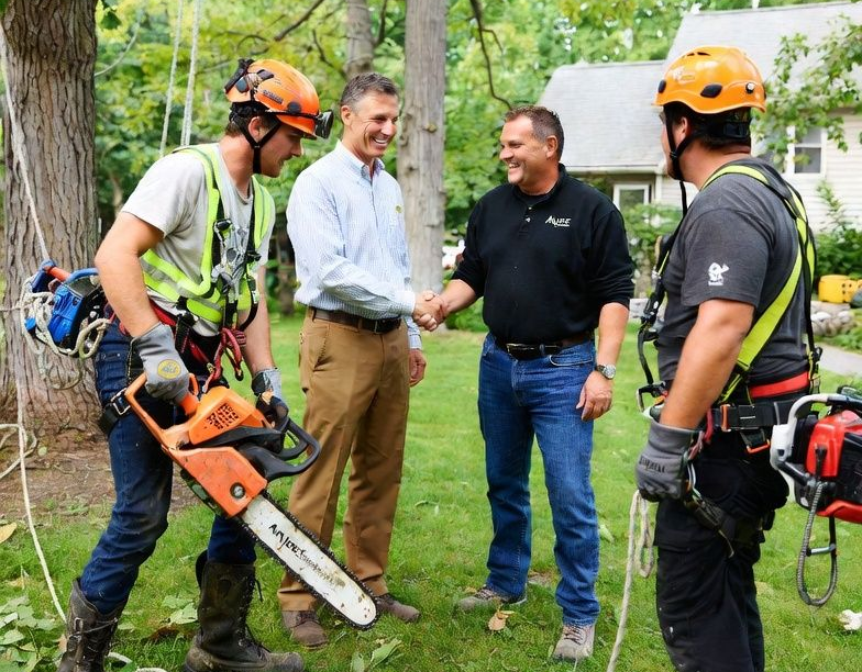 Two people in climbing gear and two people in business casual clothes shake hands outside in a park-like setting.