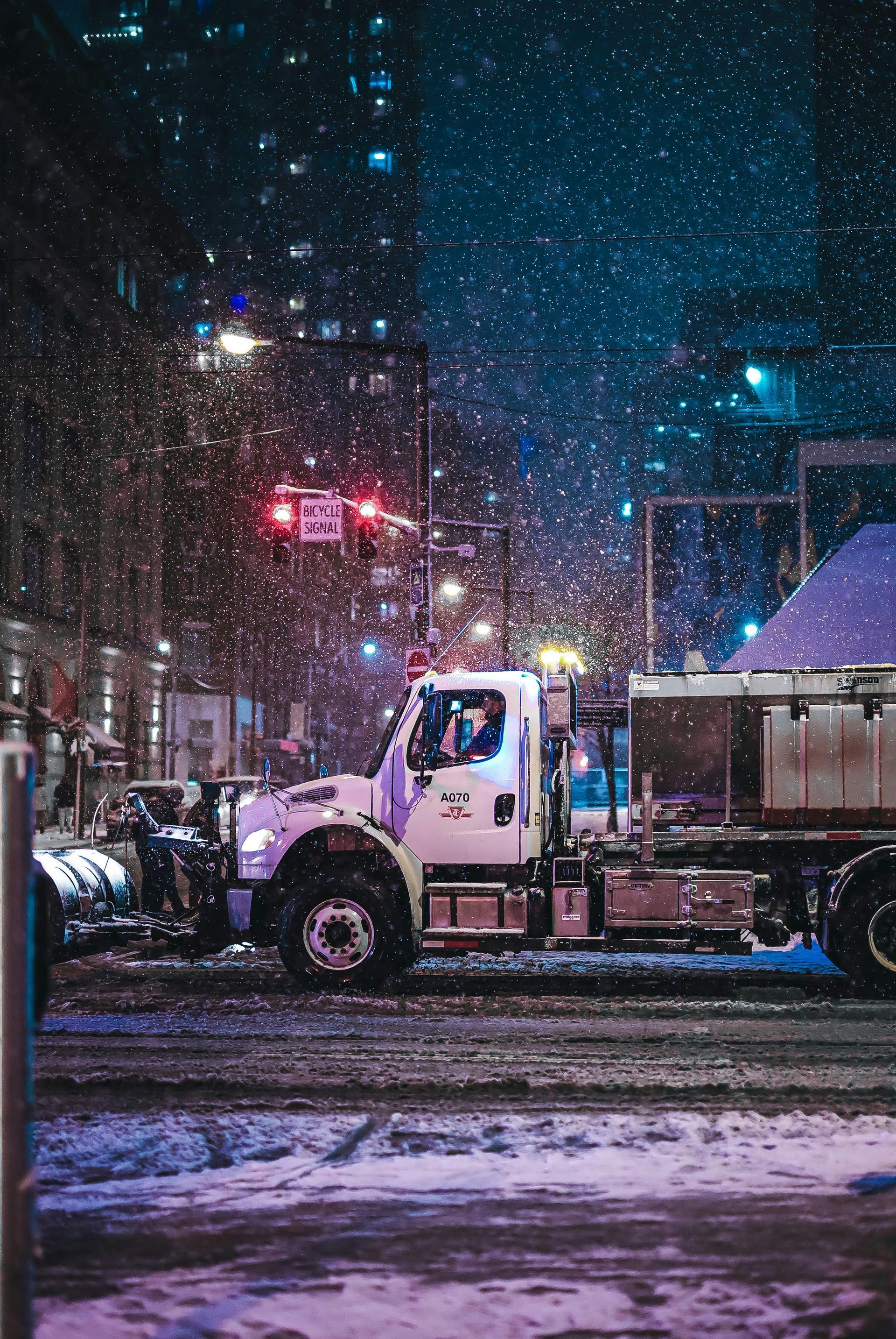 Snowplow clearing city street during snowfall at night. White truck with snow-filled bed and flashing lights.