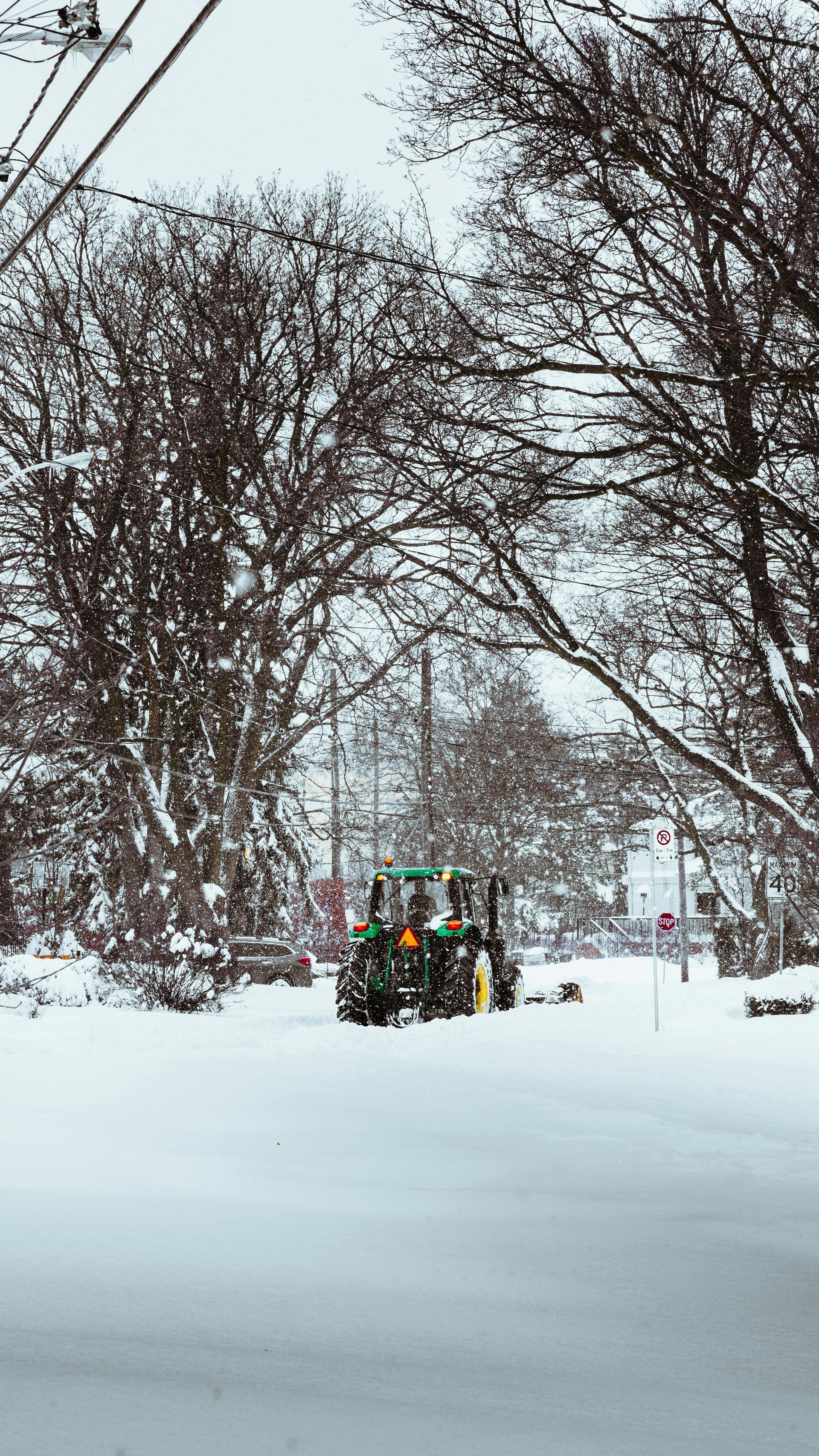 Green tractor plowing a snow-covered street between snow-dusted trees. Snowy, overcast day.