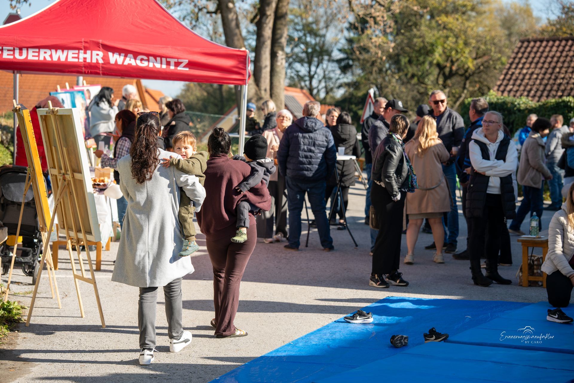 Auf einem Jahrmarkt mit einem roten Zelt, einer blauen Matte und Kindern versammelt sich eine Menschenmenge.
