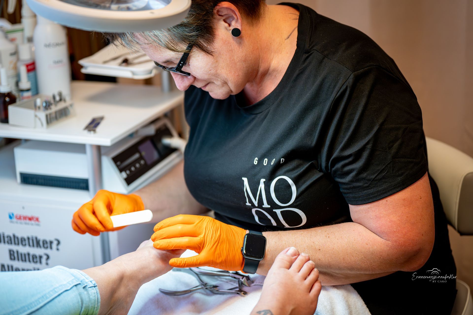 Podologe, der eine Feile am Fuß eines Patienten benutzt; orange Handschuhe, schwarzes Hemd.