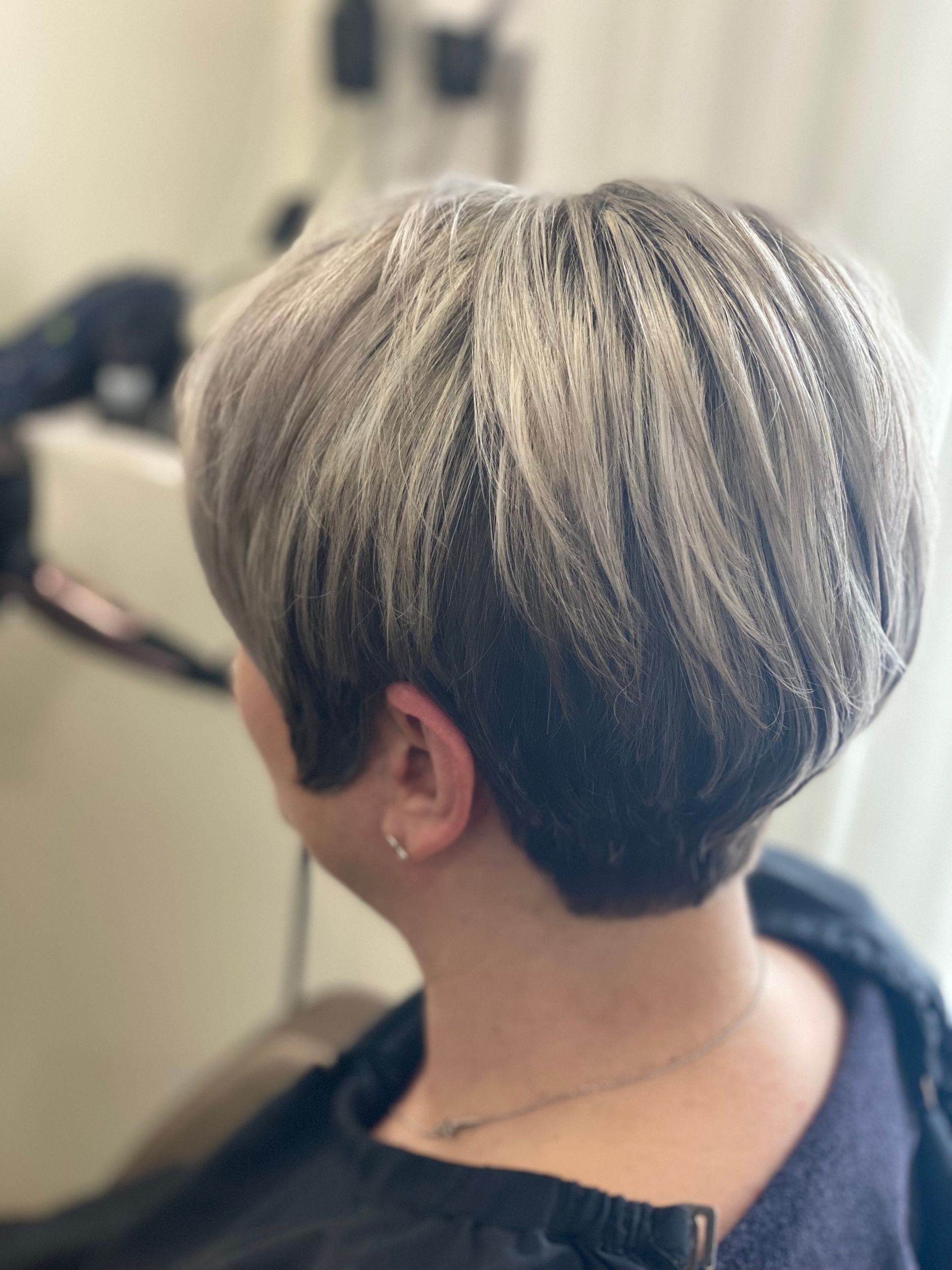 Kurzer, gestufter Pixie-Haarschnitt mit grauem Haar und dunklem Ansatz. Rückansicht einer Person im Friseursalon.