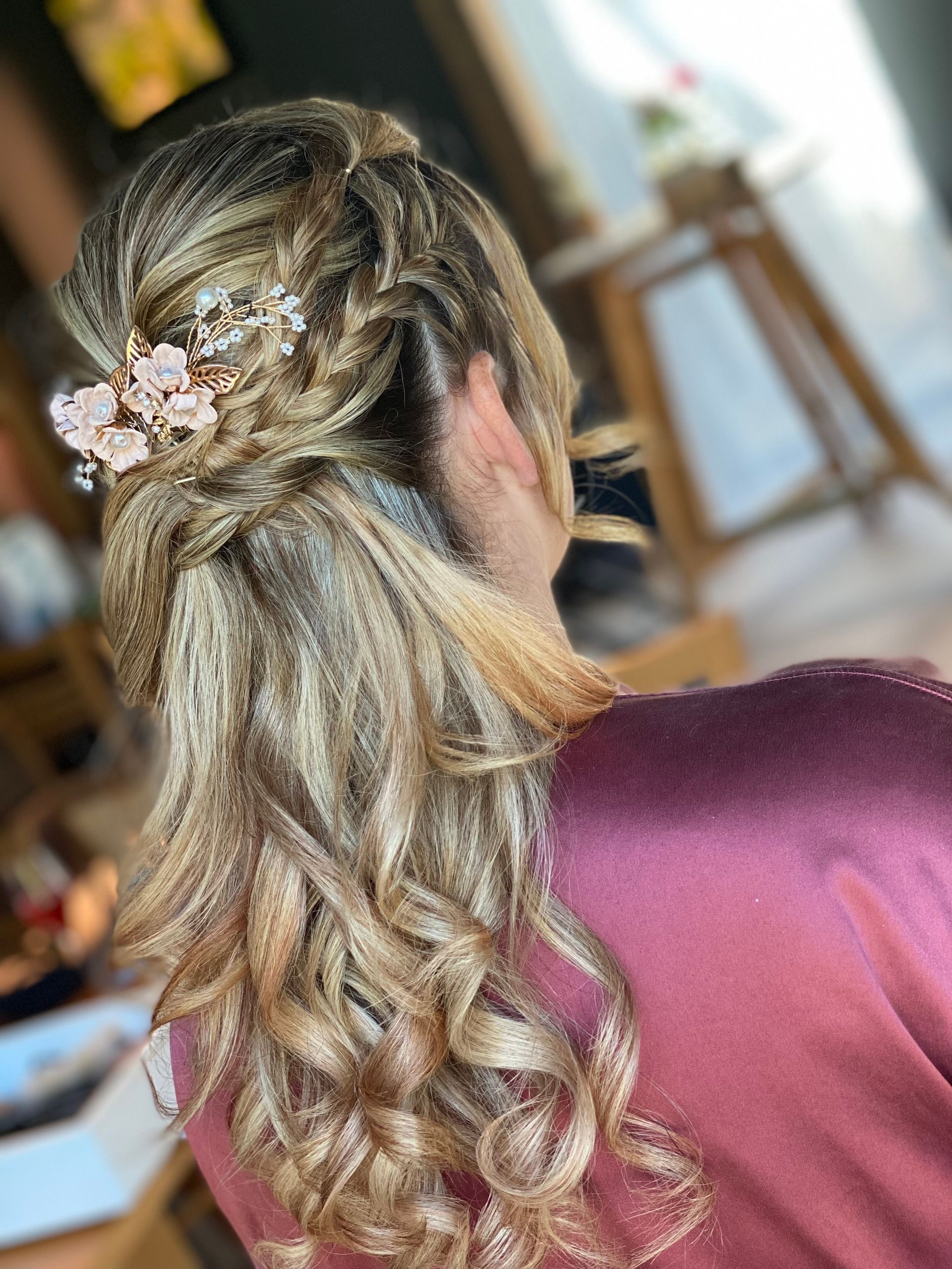 Frau mit blondem, hochgestecktem Lockenhaar und einem floralen Haarschmuck; trägt ein rosa Oberteil.