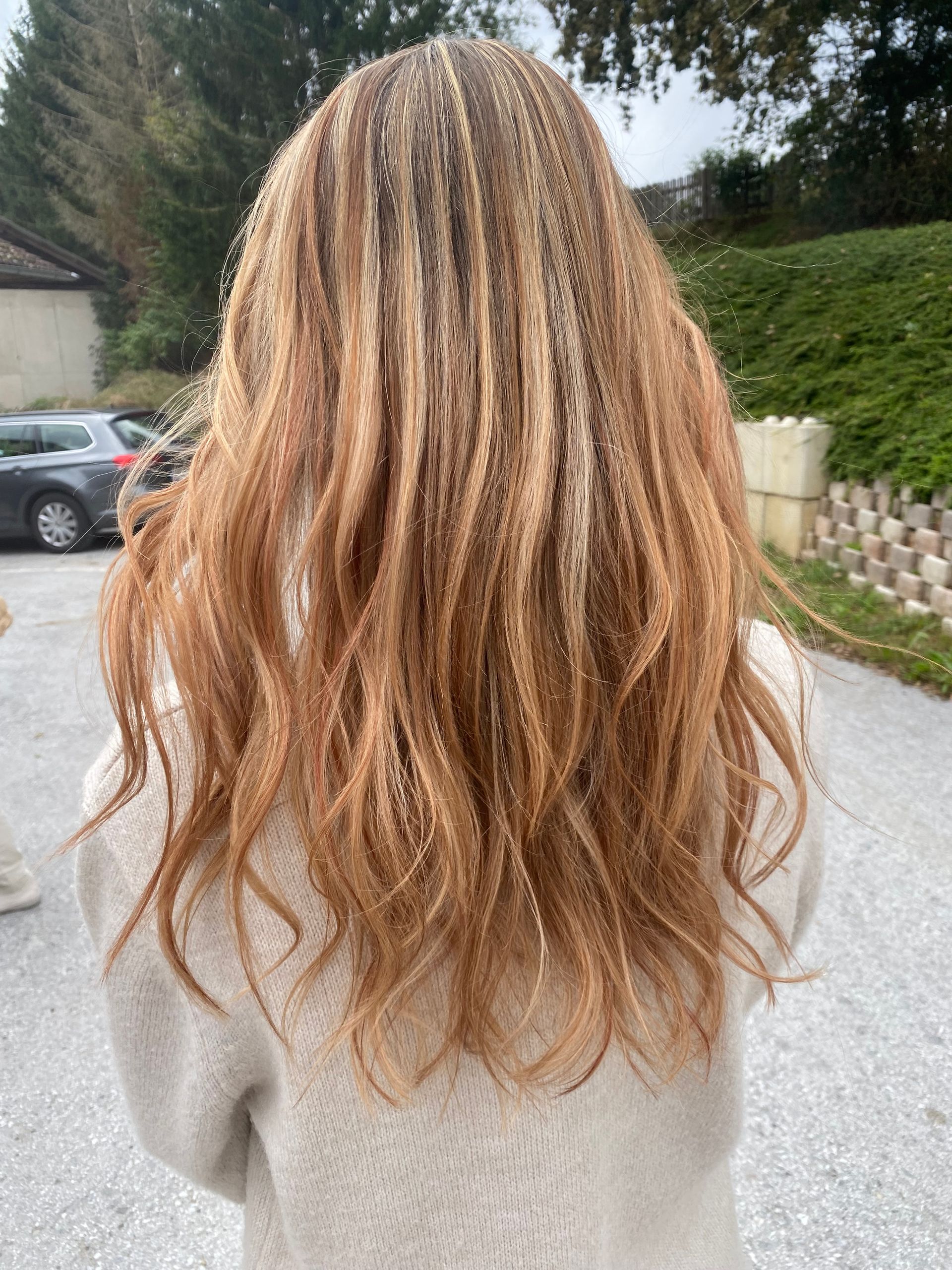 Frau mit langem, gekräuseltem, rötlich-braunem Haar, die einen beigen Pullover trägt, im Freien auf einem Steinweg.