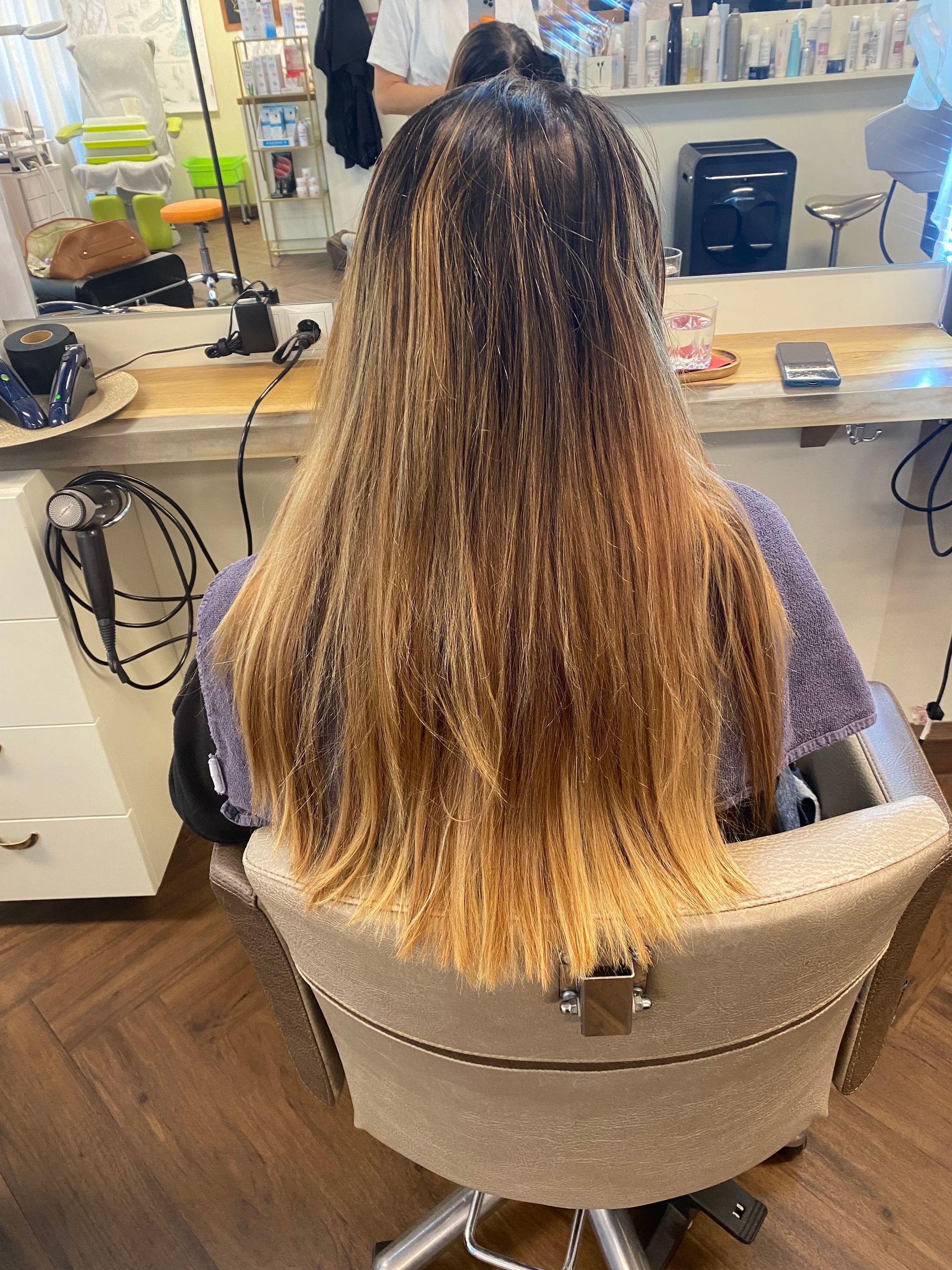 Eine Person mit langen Haaren sitzt in einem Friseurstuhl; die Haarwurzeln sind dunkel und gehen in ein Blond über.