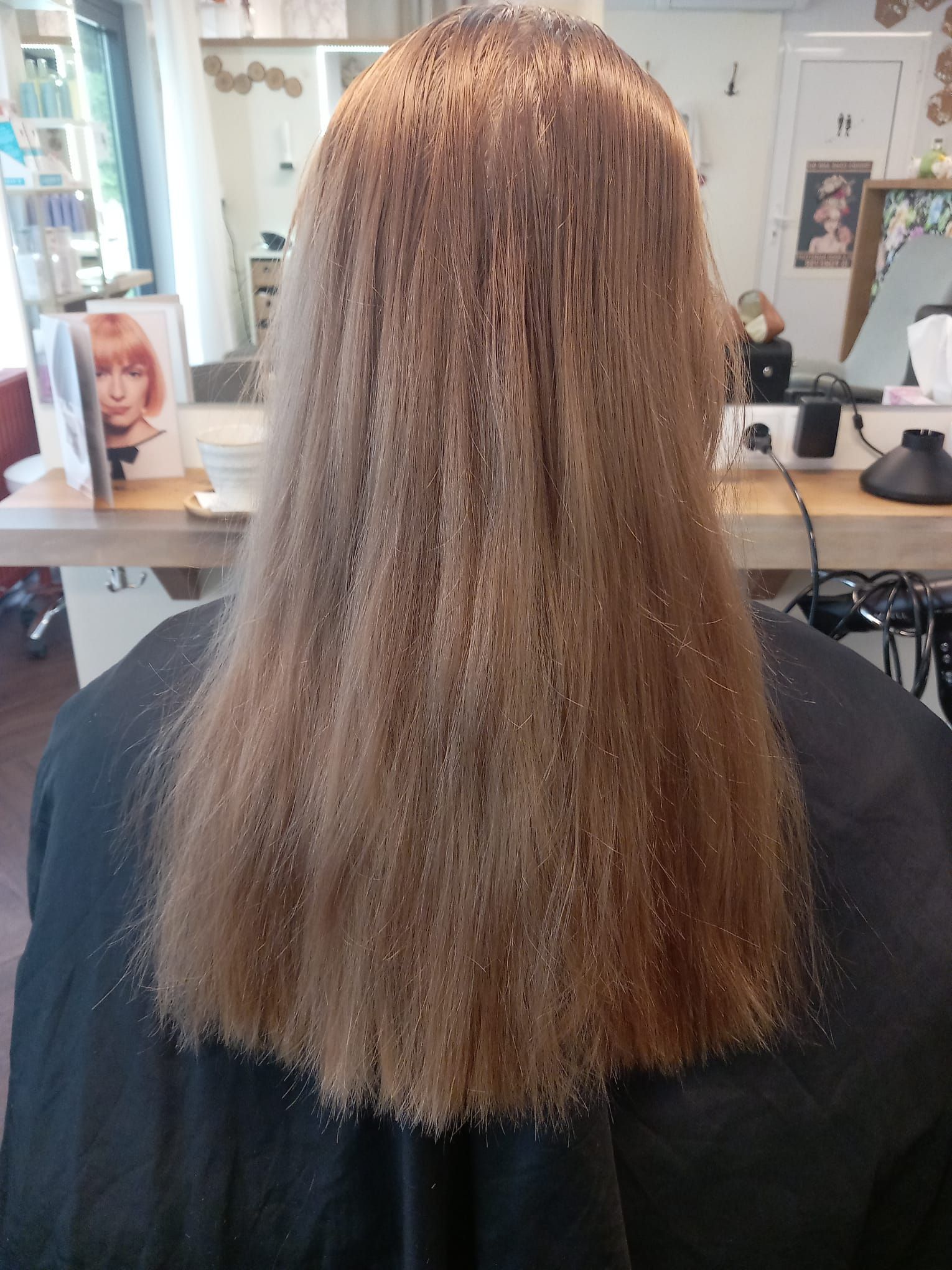 Eine Person mit langen, hellbraunen Haaren in einem Friseursalon, die einen schwarzen Umhang trägt.