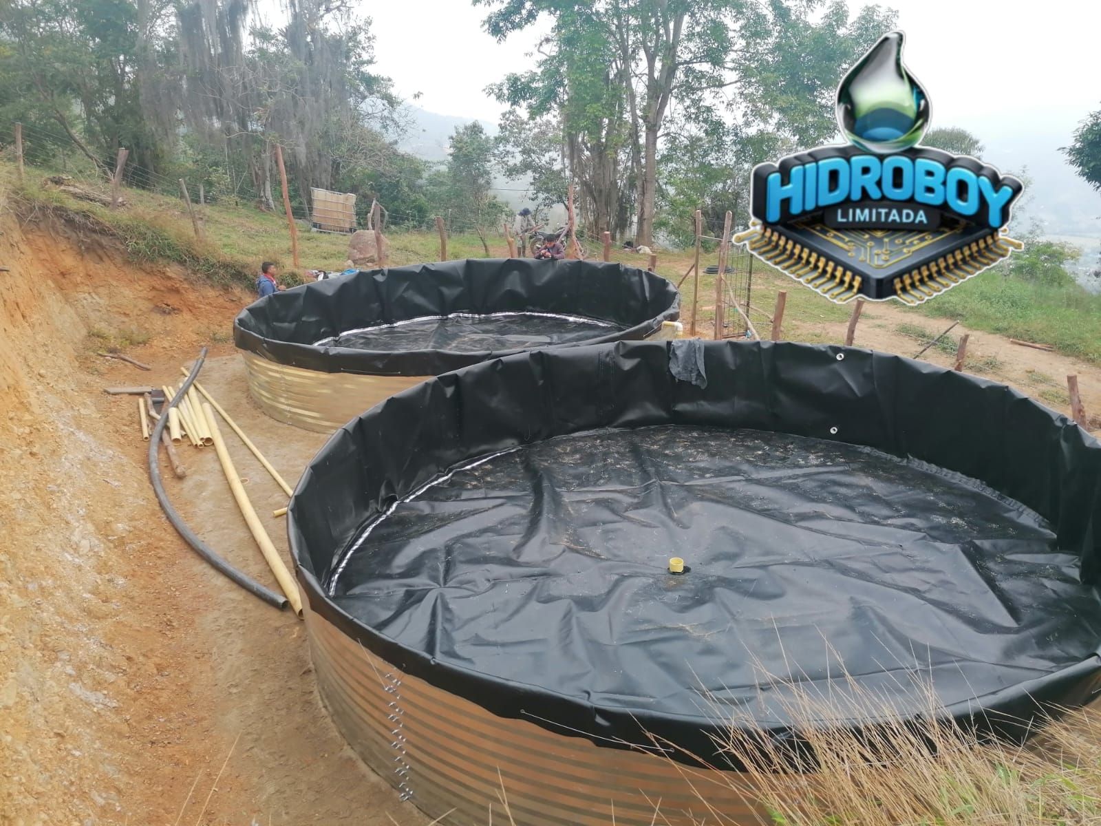 Dos grandes depósitos de agua revestidos de negro, con mangueras, en una zona rural, etiquetados como HIDROBOY LIMPEZA.