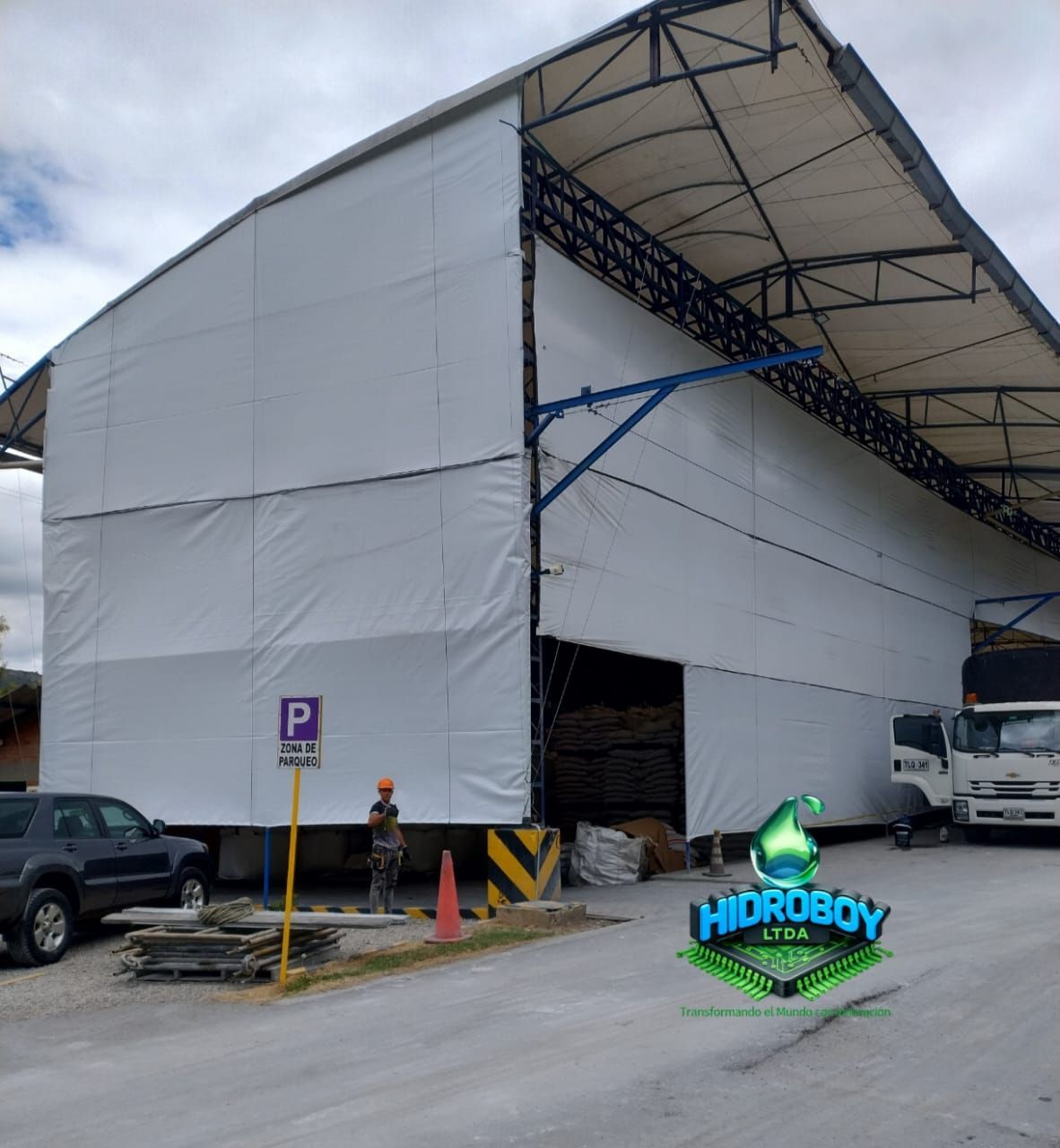Gran edificio industrial blanco en construcción con soportes de acero azul y trabajadores en la entrada.