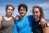 Tres personas de pie, muy juntas, al aire libre bajo un cielo azul brillante, sonriendo a la cámara.