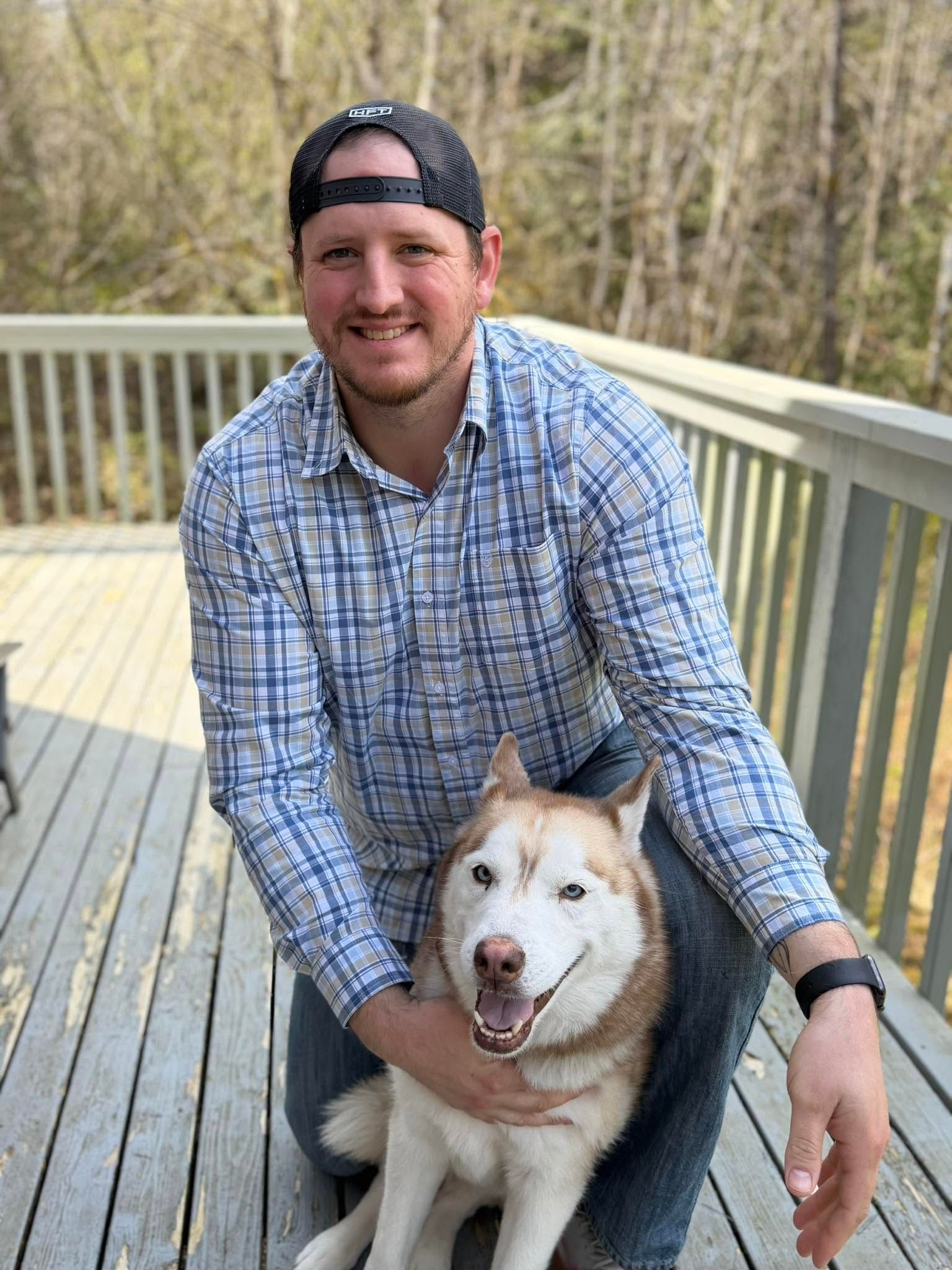 Man kneeling on a deck and hugging a smiling husky in a plaid shirt and backward cap