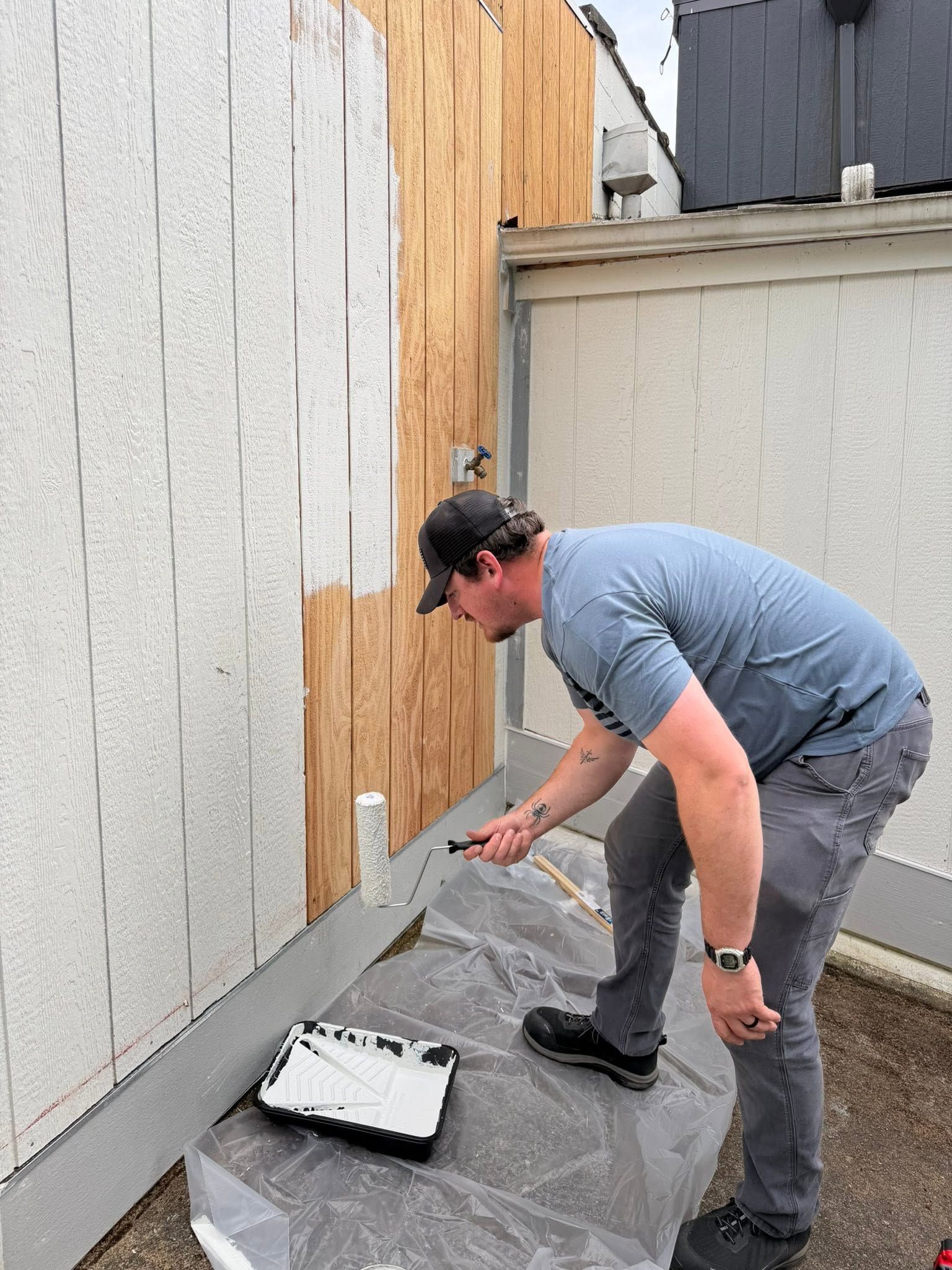 Man painting a wall with a roller beside a paint tray outdoors.