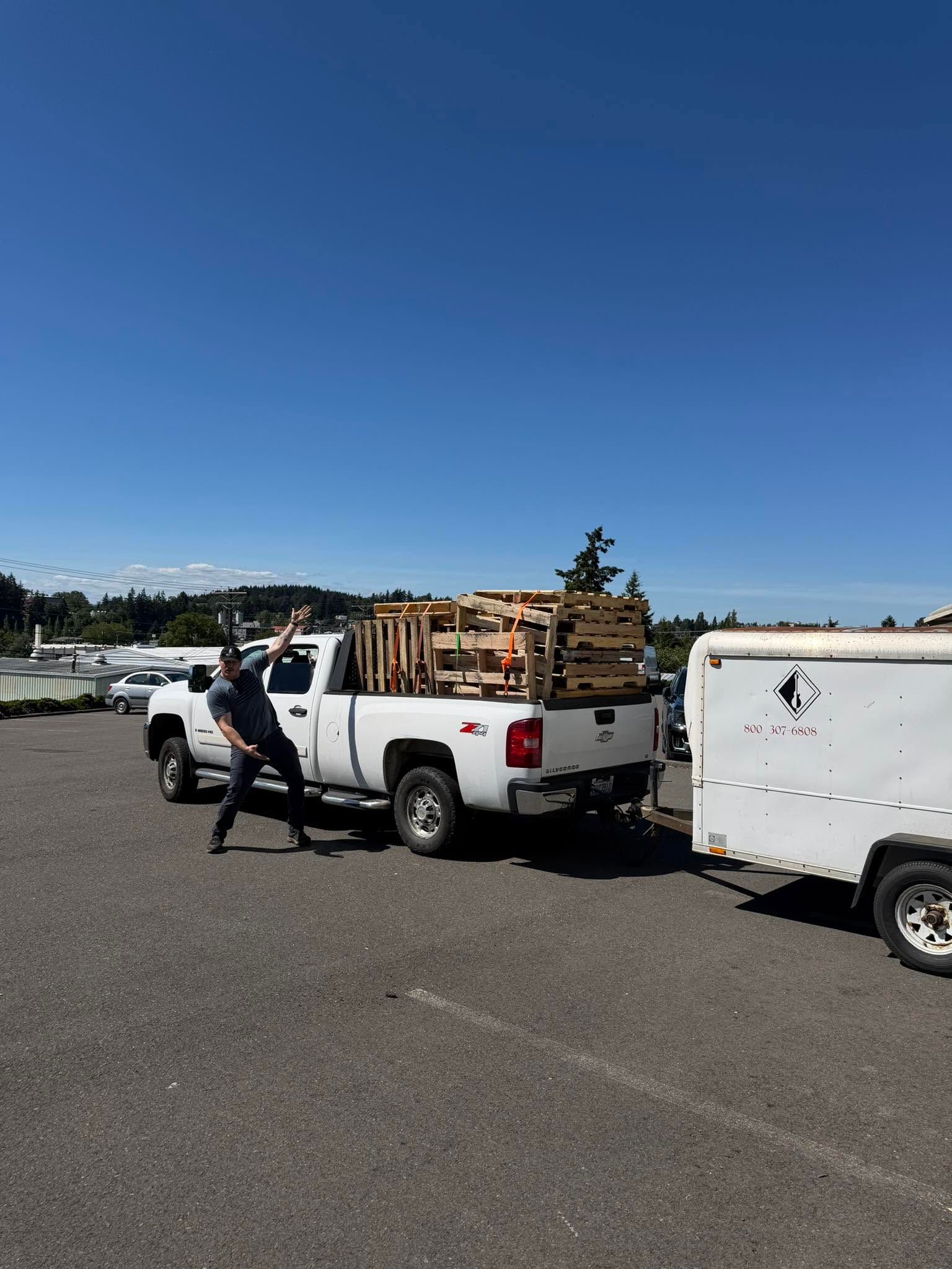 White pickup truck loaded with wood, parked beside a white trailer in a sunny open lot
