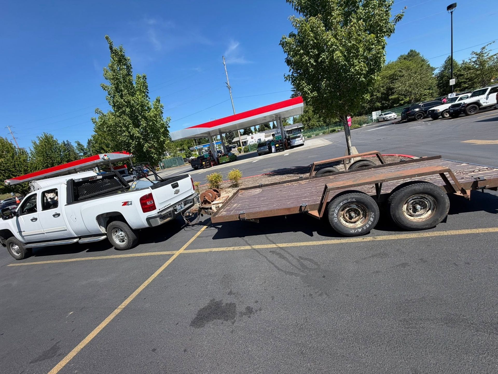 White pickup truck pulling a rusty flatbed trailer in a parking lot under a clear blue sky