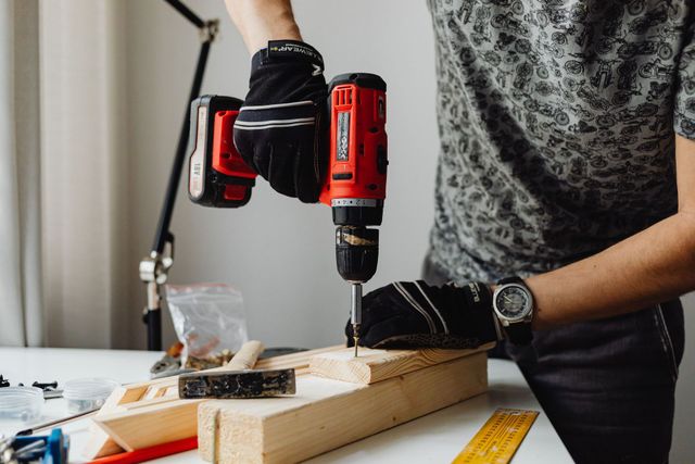 Person drilling into a wooden board on a workbench with a red cordless drill.