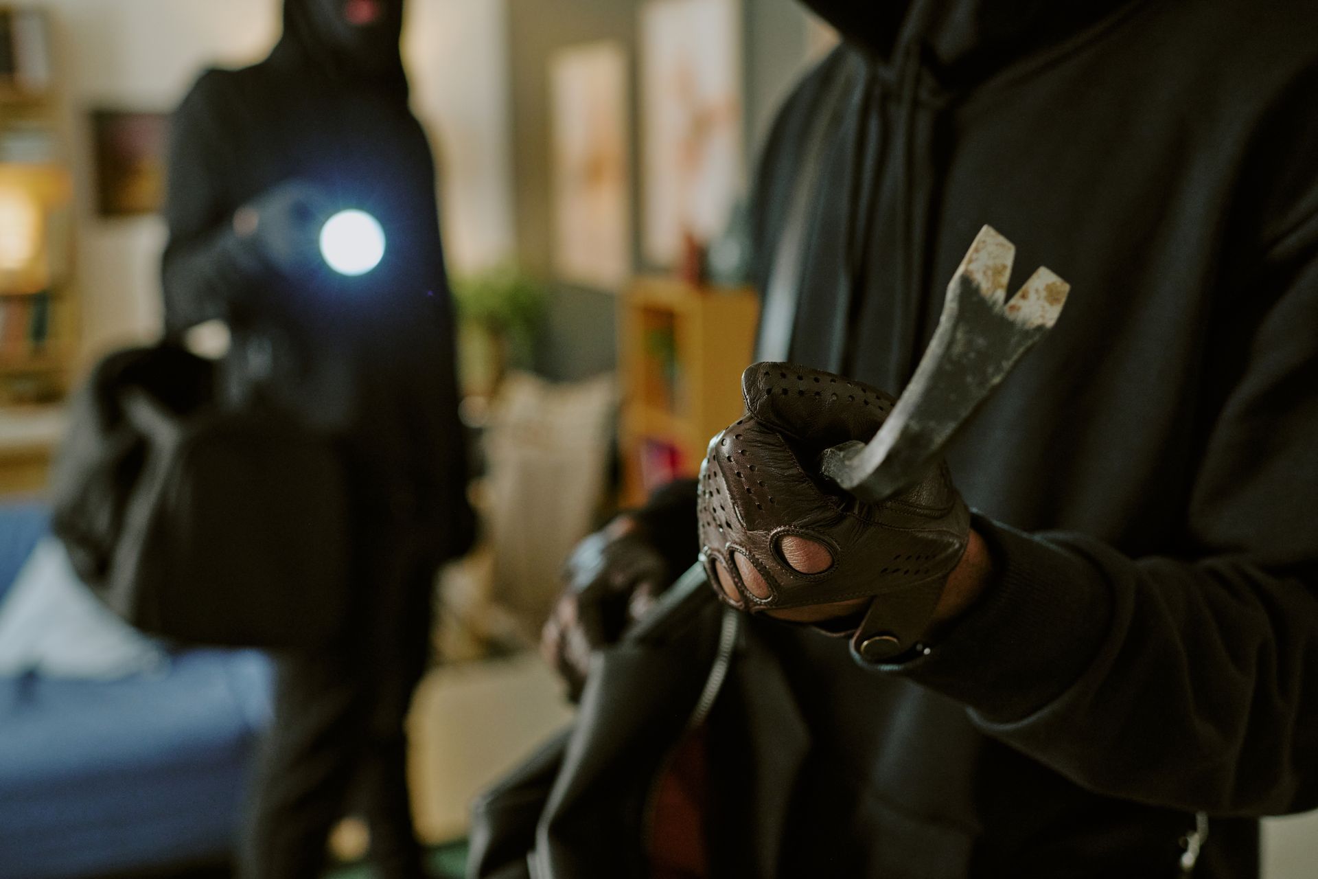Two figures, masked in black, inside a room. One holds a crowbar, other holds a flashlight and a bag.
