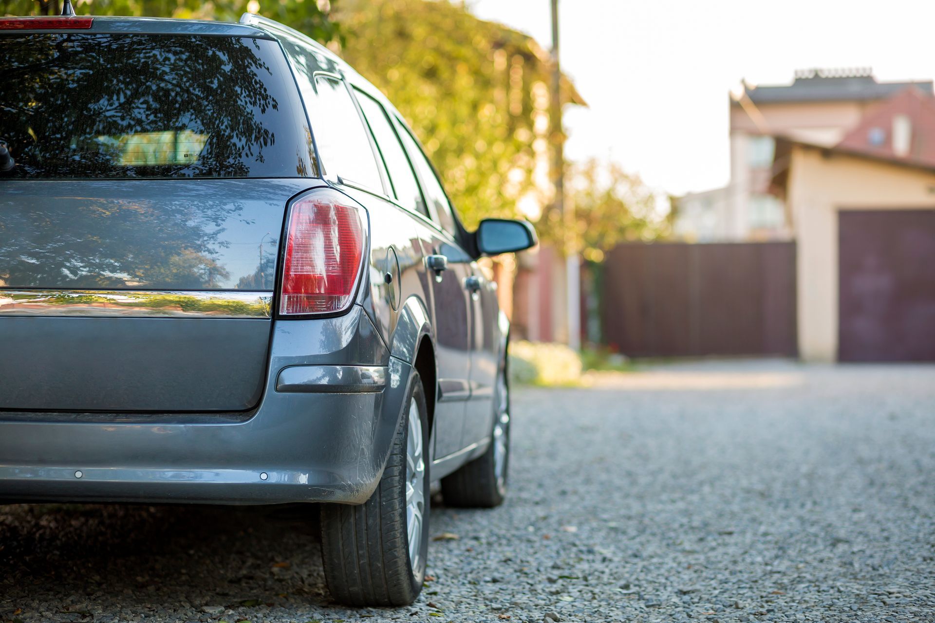 Grey station wagon parked on gravel road next to a house with a wooden fence.