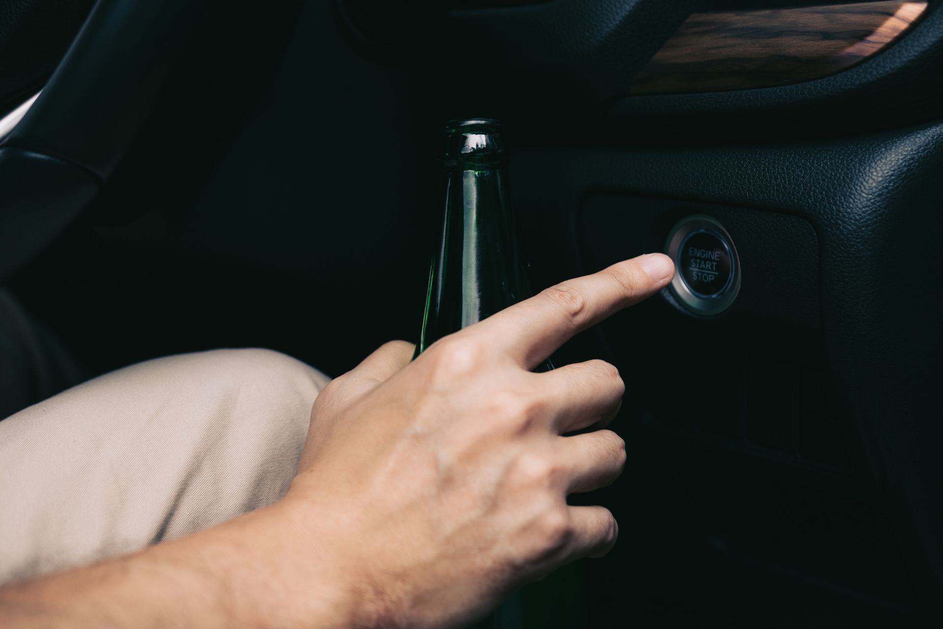 A hand holding a beer bottle reaches for a car's ignition button, inside a vehicle.