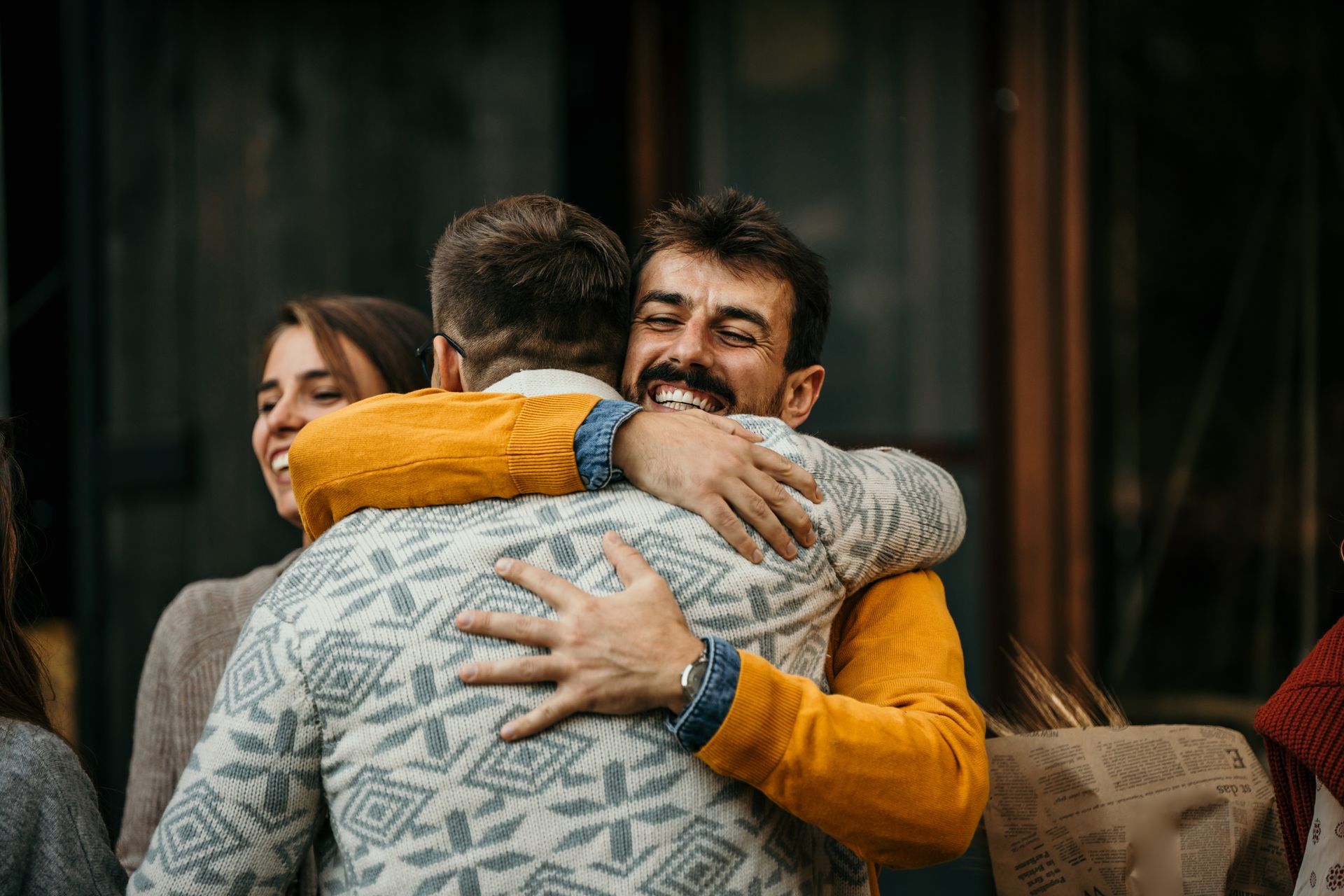 Group of friends embracing; outdoors. Man in yellow sweater smiles, hugging another man with gray sweater.